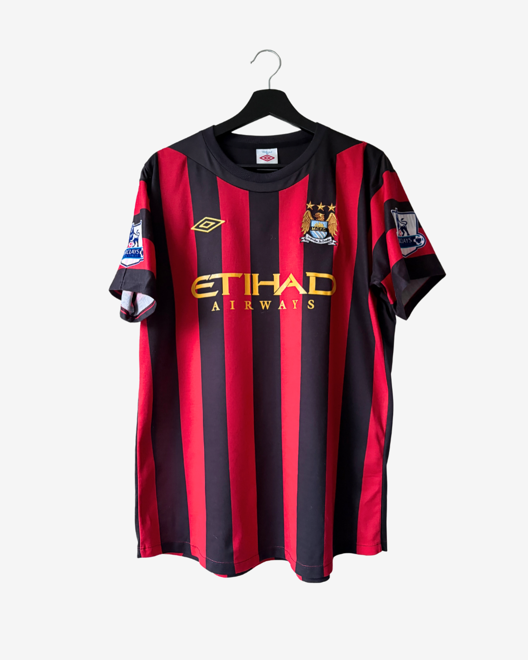 A Manchester City home soccer jersey with red and black vertical stripes, featuring the gold embroidered Umbro logo, the club crest, and 'ETIHAD AIRWAYS' sponsor text, hanging on a black hanger against a light gray background.