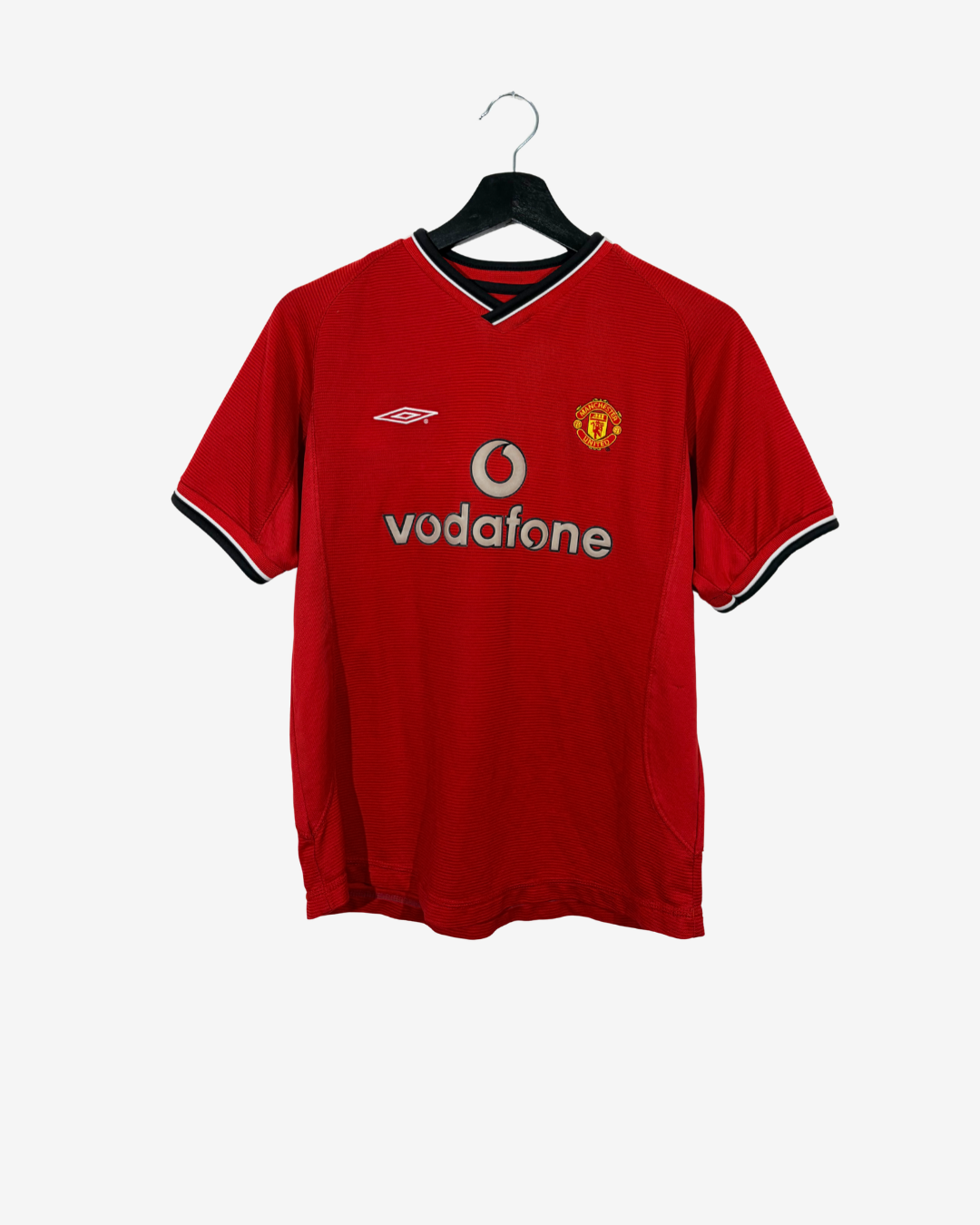 A red Manchester United short-sleeve soccer jersey with black and white trim on the collar and cuffs, featuring the silver Vodafone sponsor logo and the club crest on the chest, hanging on a black hanger against a light gray background.