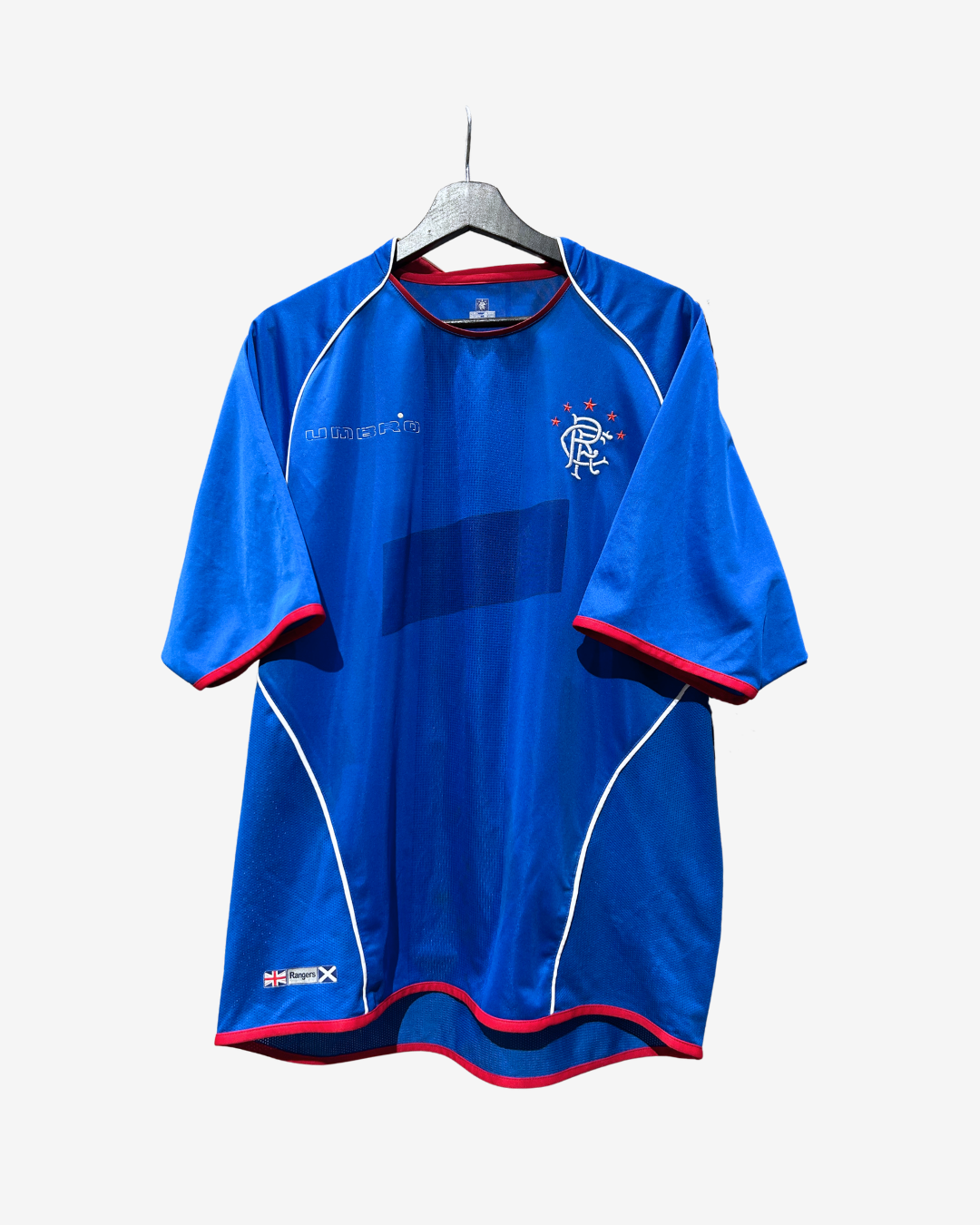 A blue short-sleeve soccer jersey with red and white trim, hanging on a black hanger against a light gray background. The jersey features the Rangers FC crest on the left chest and a sponsor logo on the right chest.