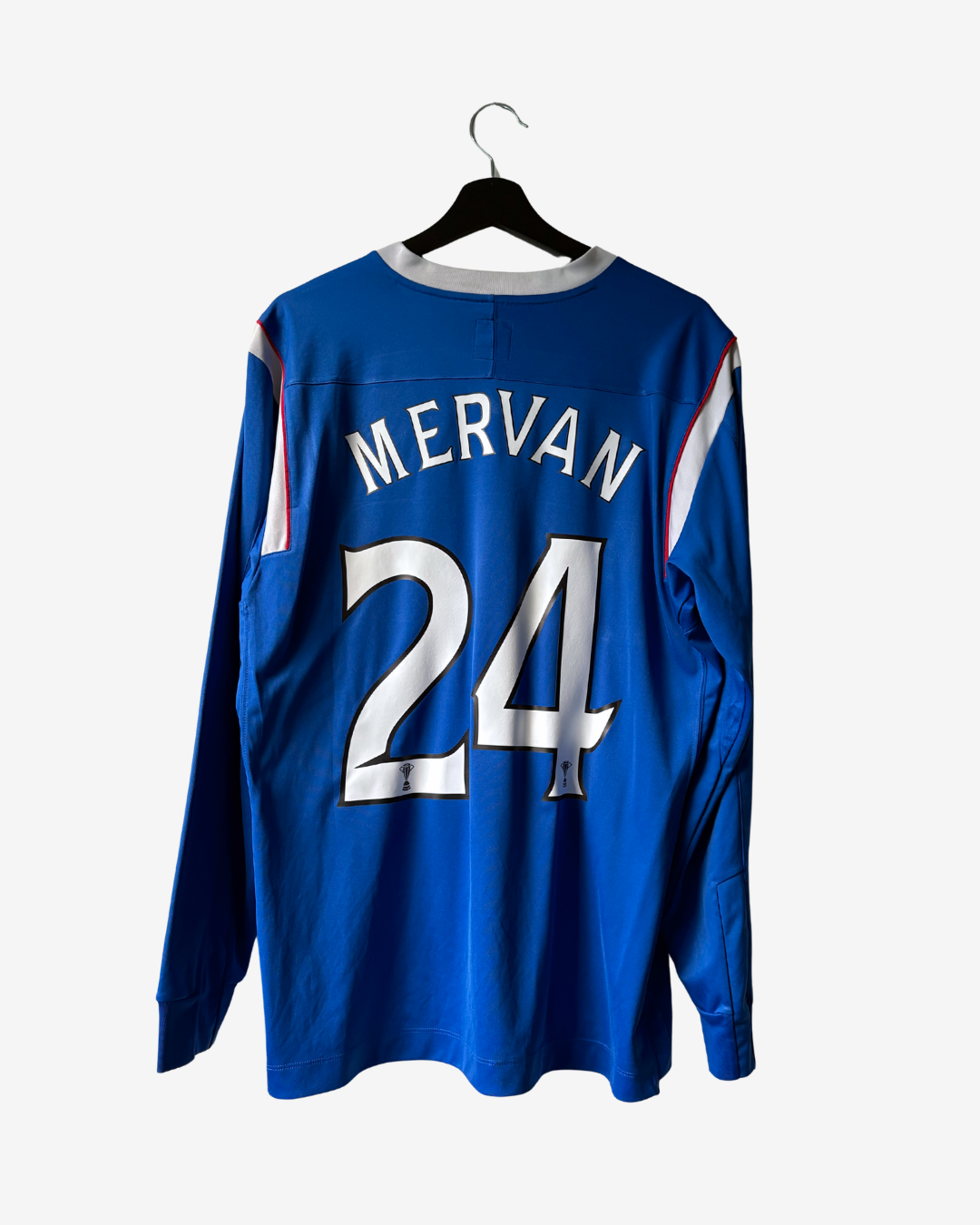 A royal blue long-sleeve soccer jersey hanging on a black hanger against a light gray background. The back of the jersey features the name 'MERVAN' and the number '24' in white with black outlines, and there are white, red, and blue stripes on the shoulders.