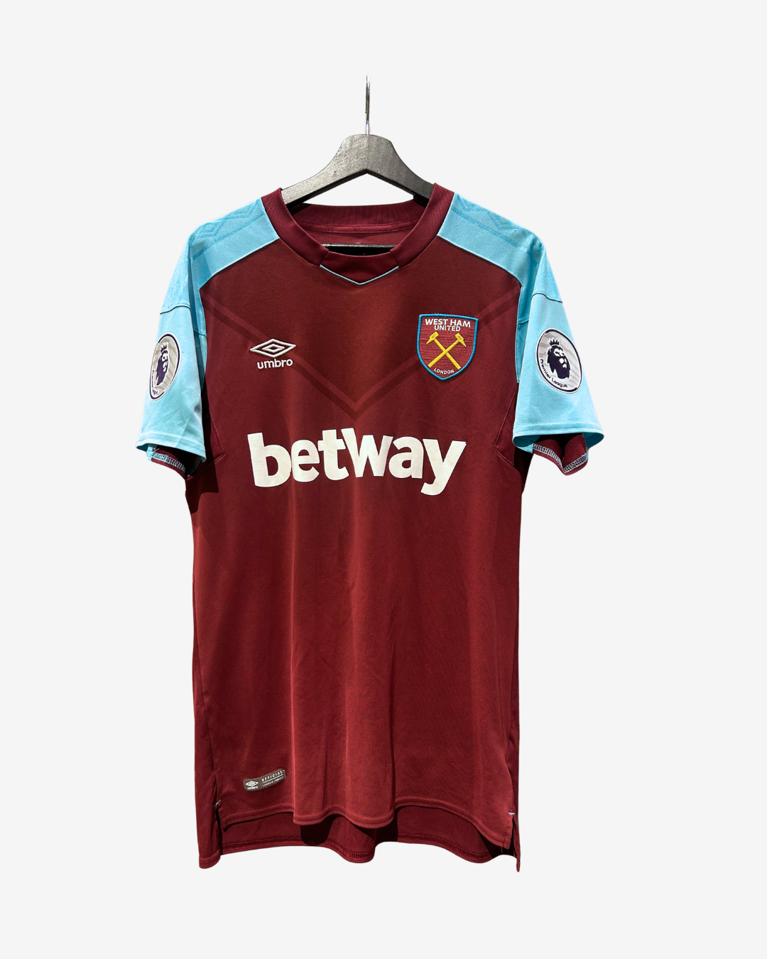 A West Ham United home soccer jersey by Umbro, featuring a maroon body, light blue shoulder panels, and a white 'betway' sponsor logo on the chest, hanging on a wooden hanger against a light gray background.