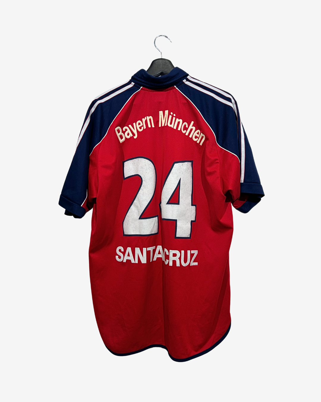A red and navy blue Bayern München soccer jersey with the name 'SANTACRUZ' and number '24' printed on the back, hanging on a black hanger against a light gray background.