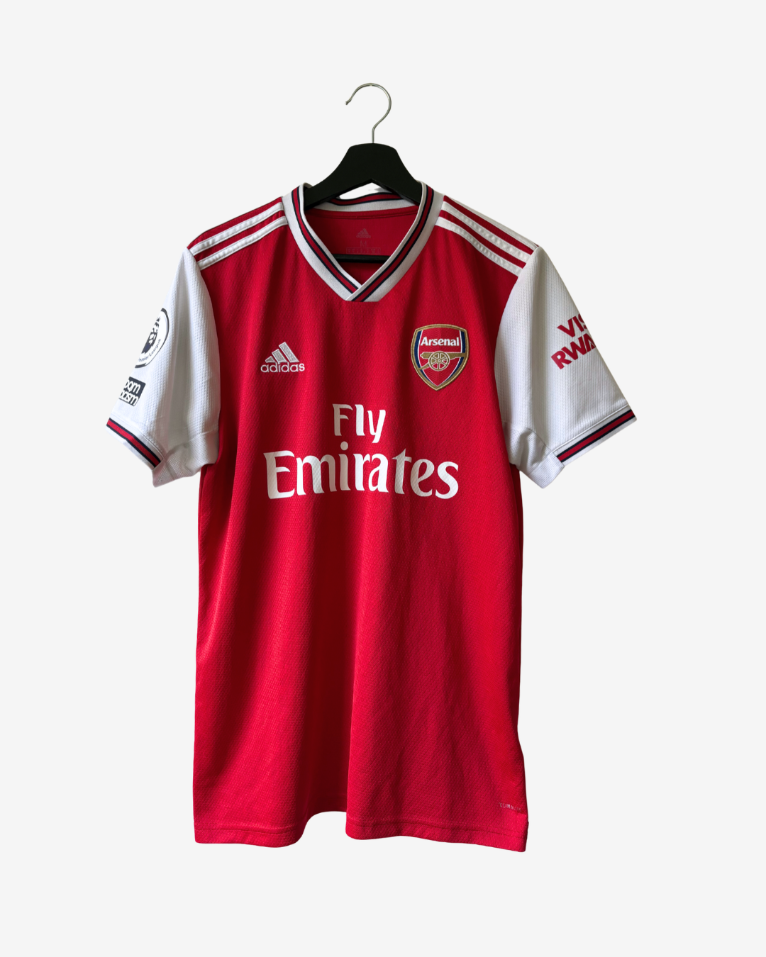A red and white Arsenal home soccer jersey by Adidas, hanging on a black hanger against a light gray background. The jersey features the Arsenal club crest, the Adidas logo, and the Fly Emirates sponsor logo.