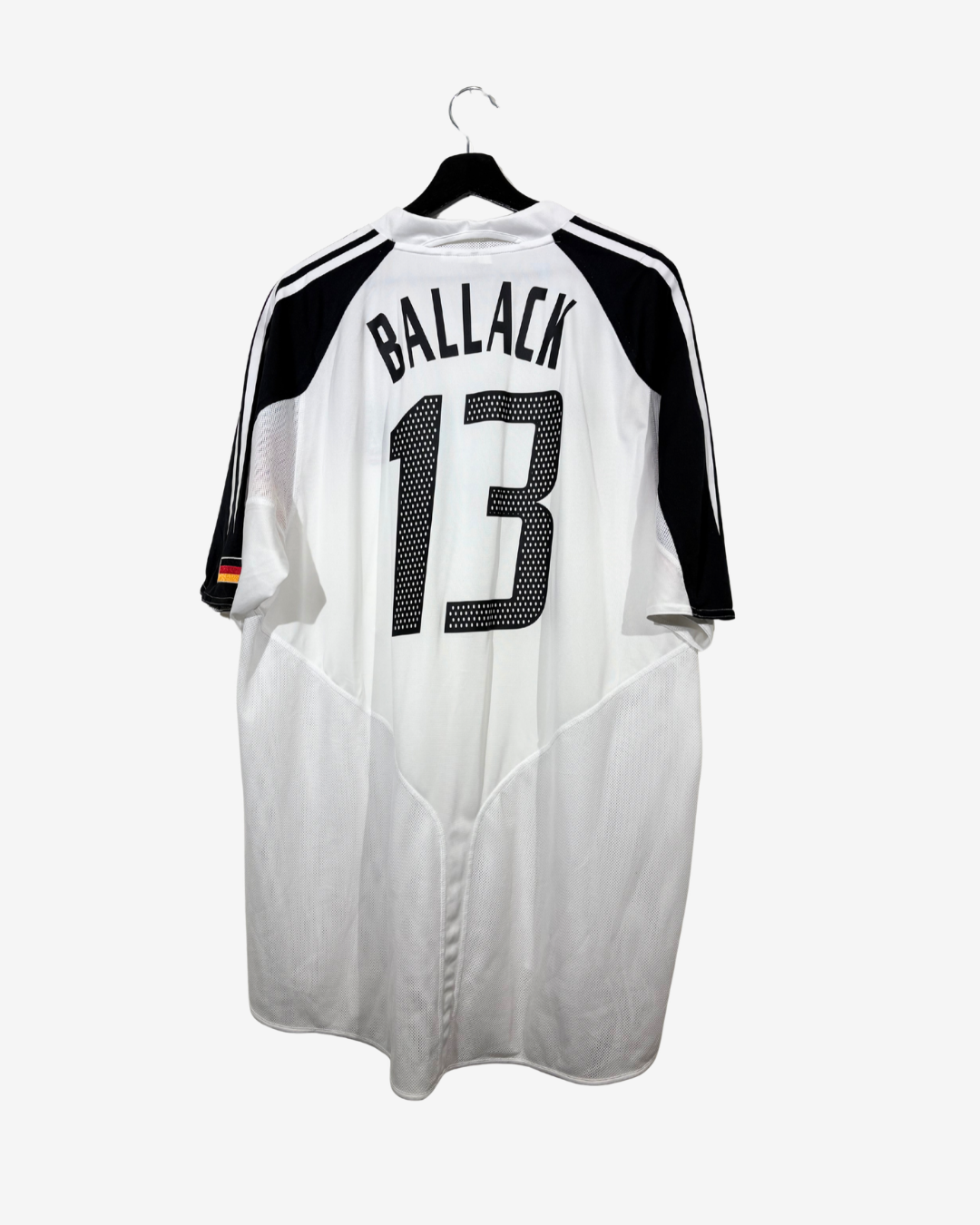 A white soccer jersey with black shoulder panels, black stripes, and 'BALLACK 13' printed on the back, hanging on a black hanger against a light gray background.