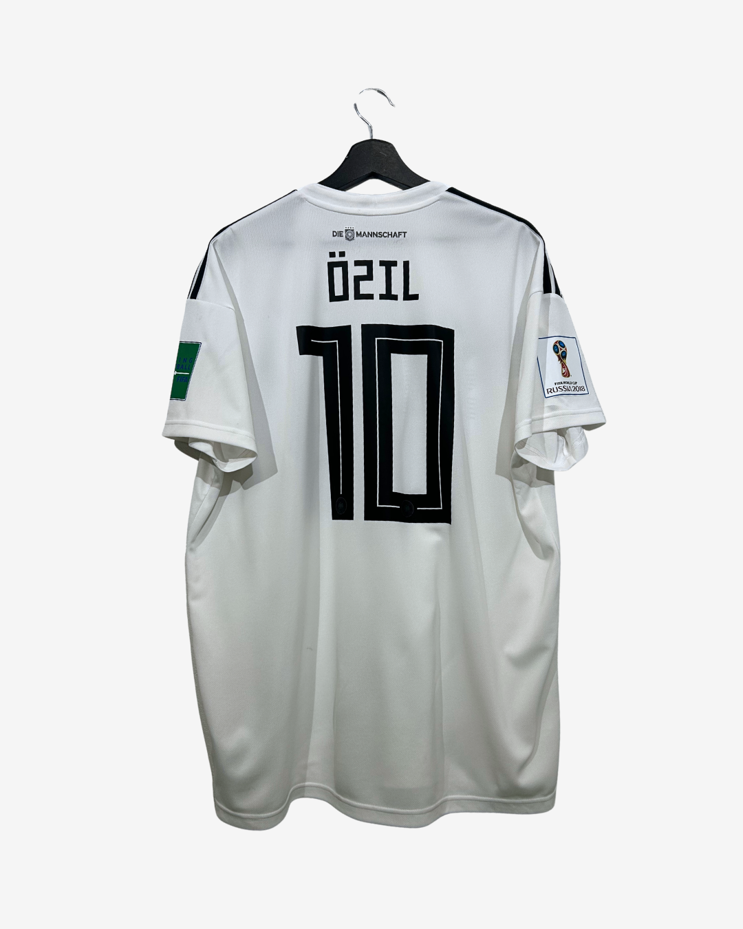A white Germany national team soccer jersey with 'ÖZIL' and the number '10' printed in black on the back, hanging on a black hanger against a light gray background.