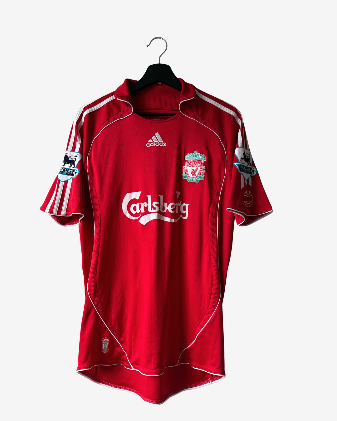 A red Adidas Liverpool soccer jersey with white stripes and Carlsberg sponsor logo, hanging on a black hanger against a light gray background.