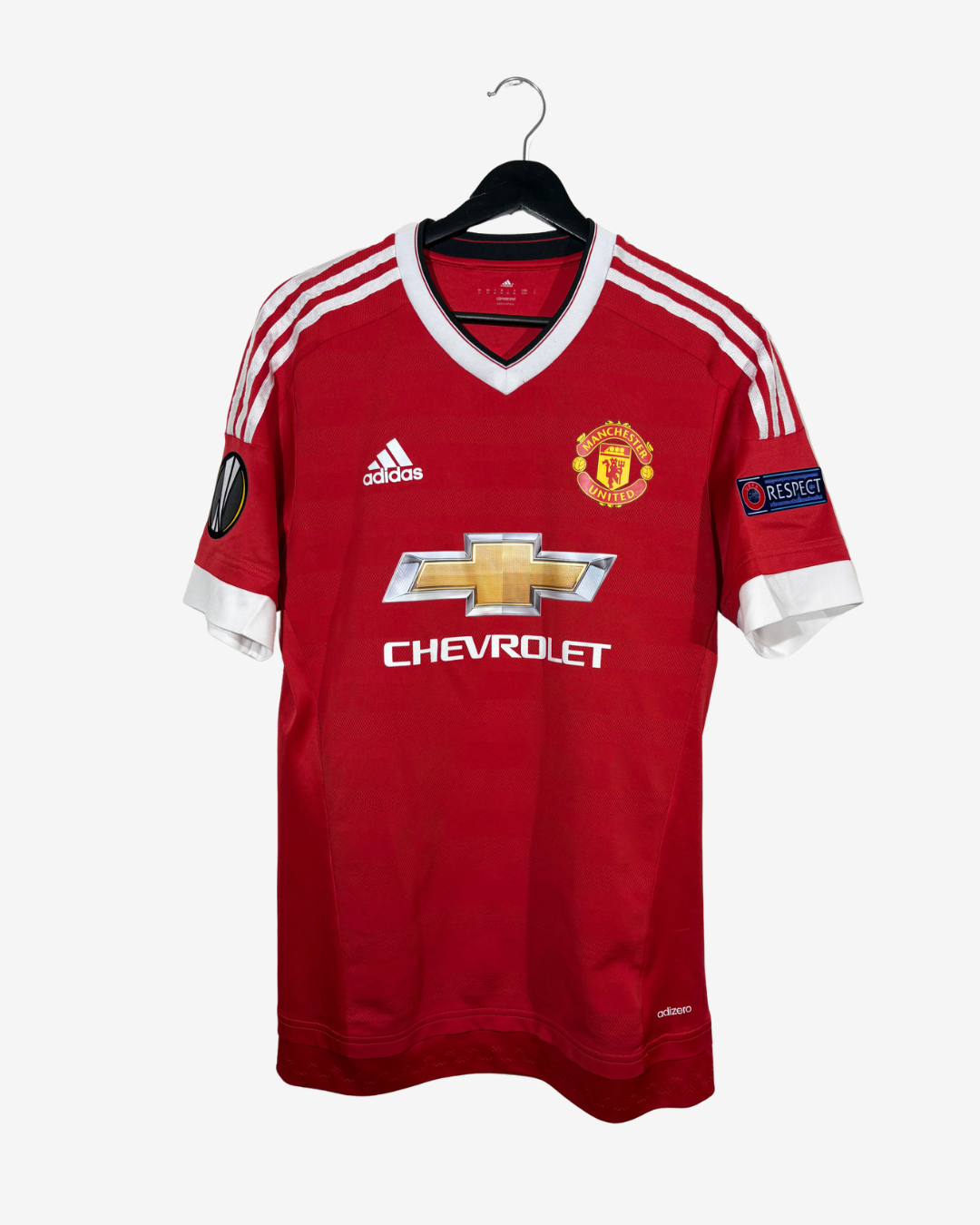 A red Manchester United Adidas soccer jersey with white shoulder stripes, a white V-neck collar, and a gold Chevrolet logo on the front, hanging on a black hanger against a light gray background.