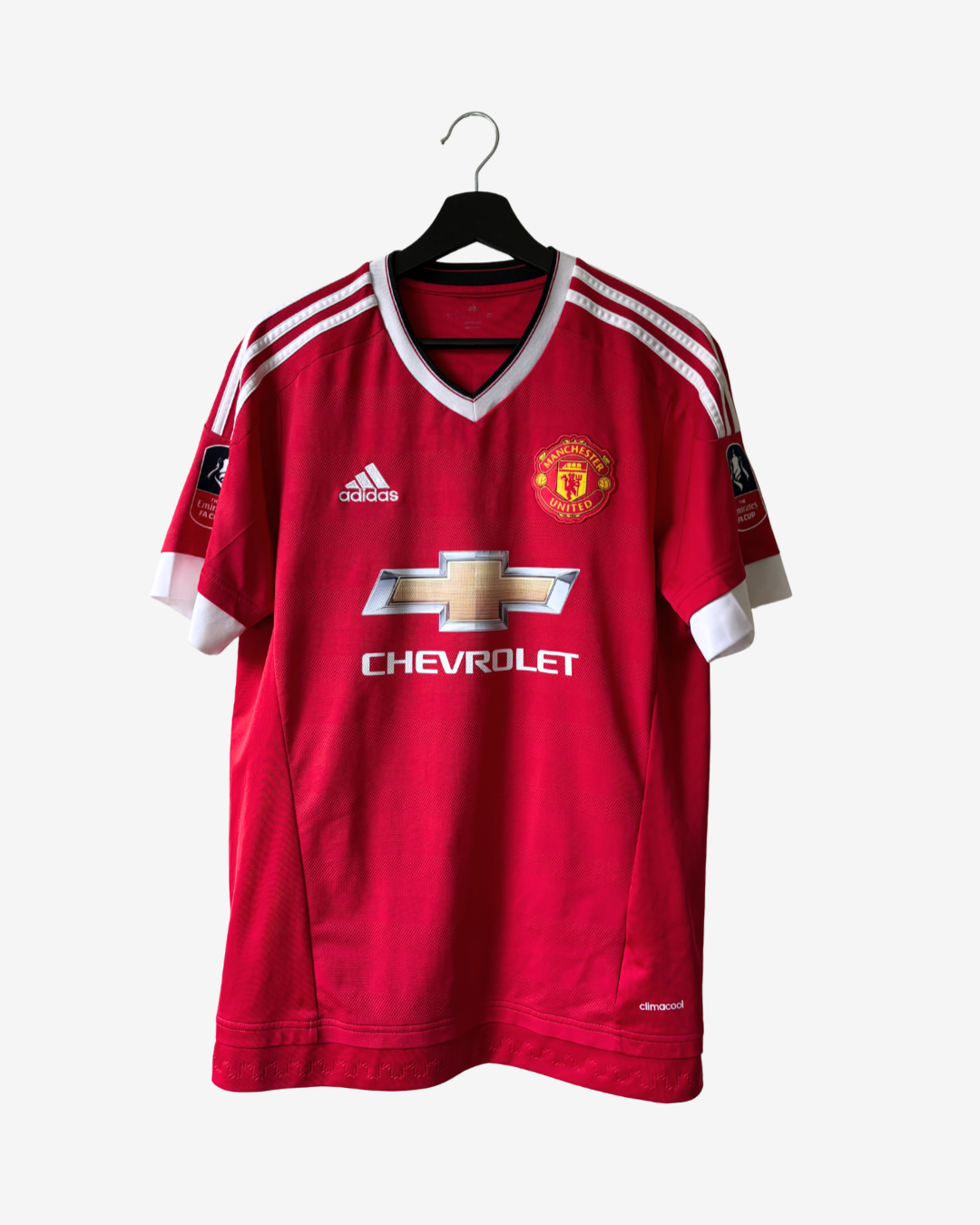 A red Manchester United soccer jersey by Adidas, hanging on a black hanger against a light gray background. The jersey features white shoulder stripes, a white V-neck collar, the Manchester United crest on the right chest, the Adidas logo on the left chest, and the Chevrolet sponsor logo across the center.