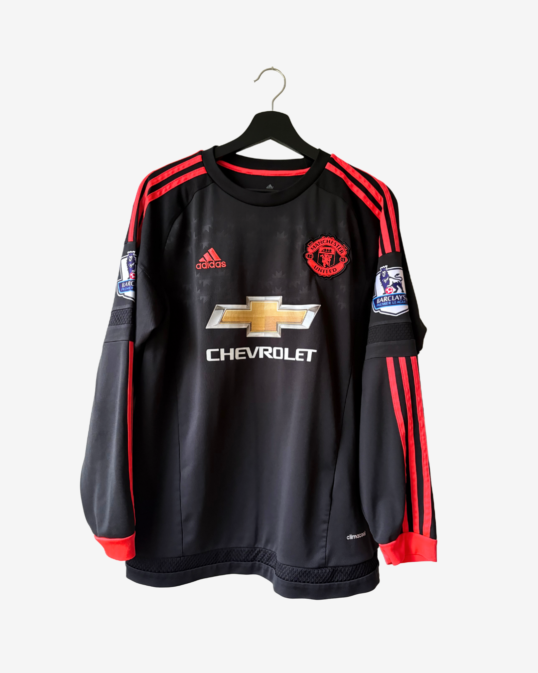A black long-sleeve Manchester United football jersey with red stripes and a gold Chevrolet logo, hanging on a black hanger against a light gray background.