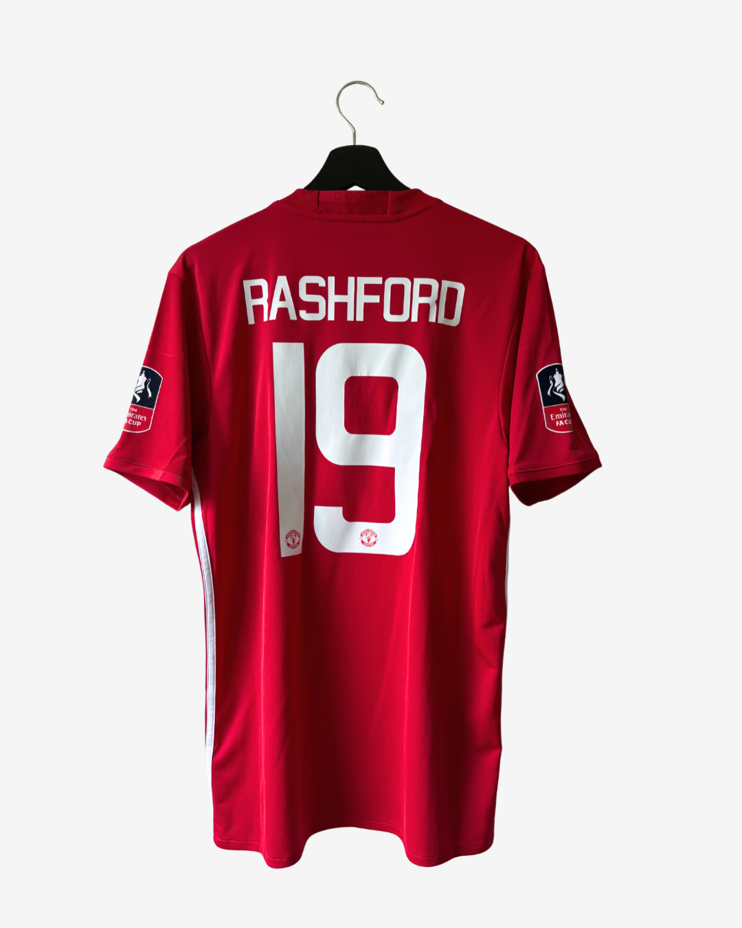A red Manchester United football jersey with 'RASHFORD' and the number '19' printed in white on the back, hanging on a black hanger against a light gray background.