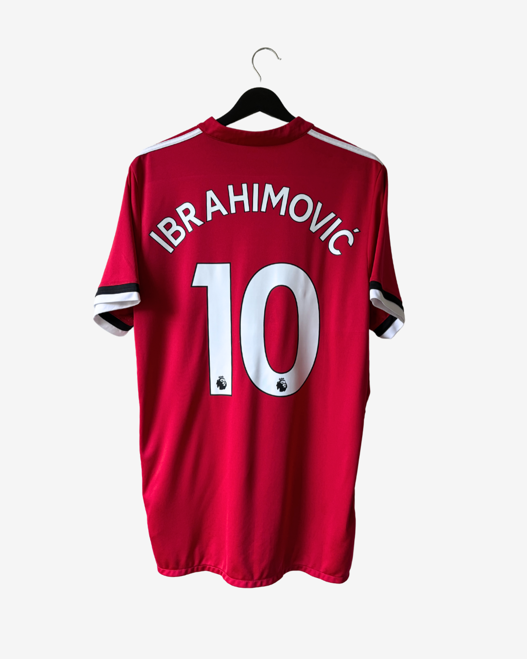 A red short-sleeve football jersey hanging on a black hanger against a light gray background. The back of the shirt features the name 'IBRAHIMOVIC' and the number '10' in white, with white shoulder stripes and black-and-white striped sleeve cuffs.