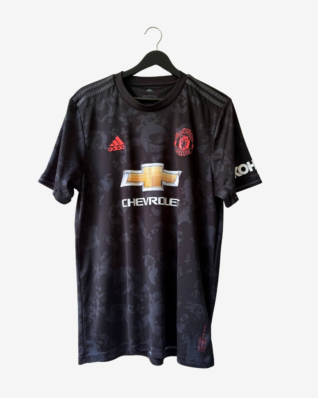 A black Manchester United short-sleeve soccer jersey with a subtle tonal camouflage pattern, featuring the red Adidas logo, Manchester United crest, and gold and white Chevrolet sponsor logo on the front, hanging on a black hanger against a light gray background.