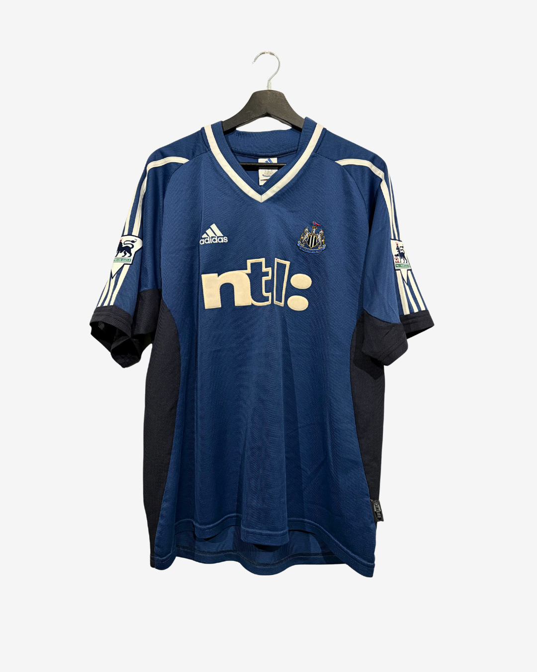 A blue Adidas Newcastle United soccer jersey with cream and black accents, hanging on a wooden hanger against a light gray background.