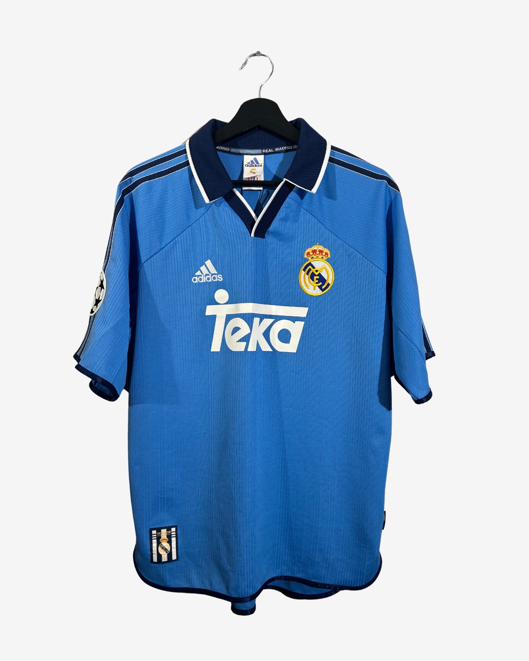 A blue Real Madrid soccer jersey by adidas, featuring the Teka sponsor logo on the front, hanging on a black hanger against a light gray background.