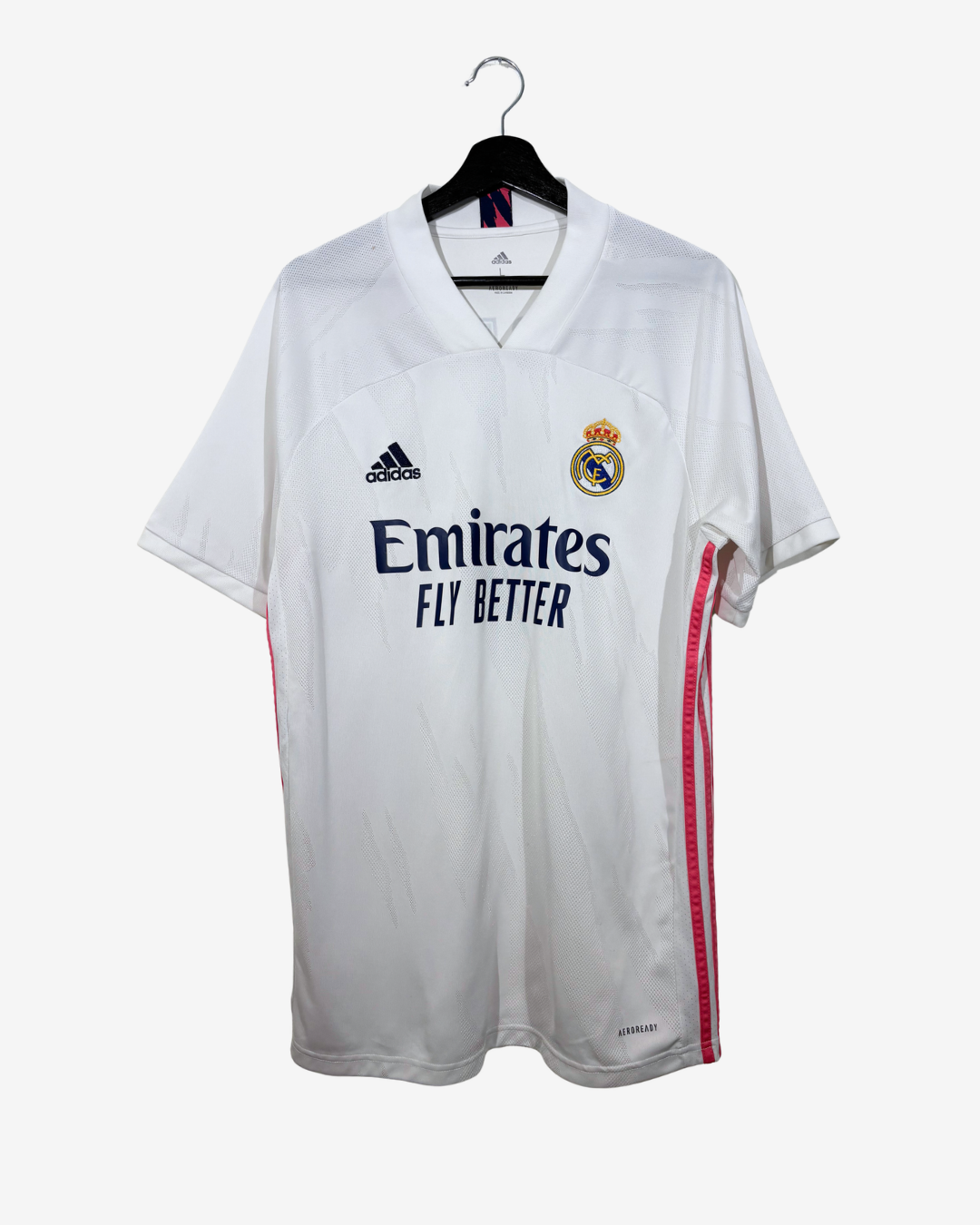 A white Real Madrid soccer jersey by adidas, featuring the Emirates Fly Better sponsor logo, hanging on a black hanger against a light gray background.