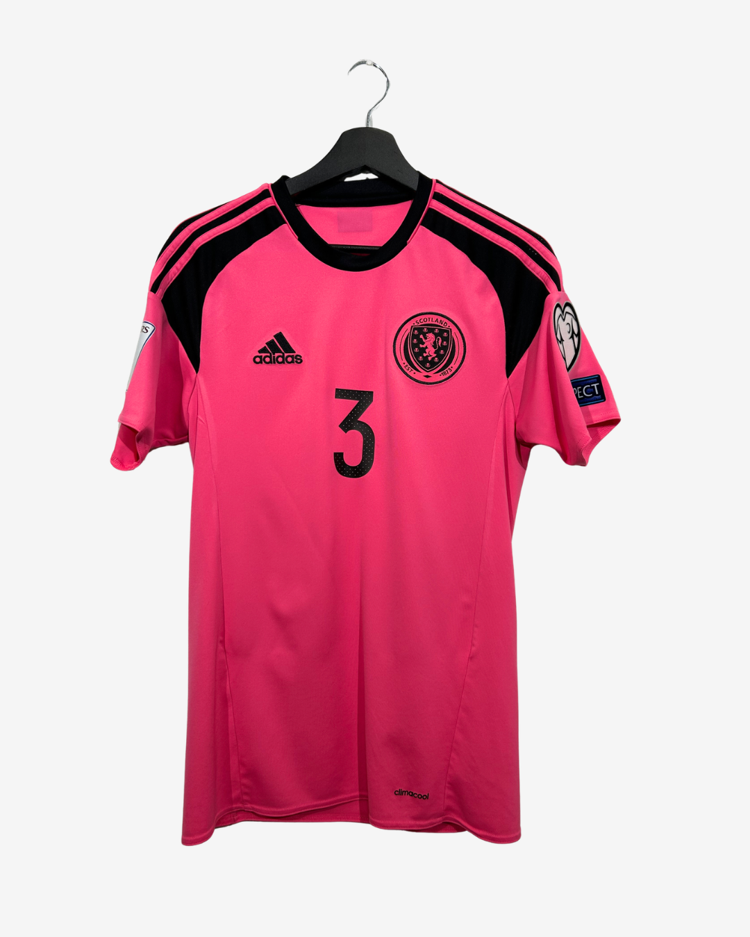 A bright pink Adidas soccer jersey with black shoulder stripes and black trim, featuring the number 3 on the chest and the Scotland national team crest. The jersey is hanging on a black hanger against a light gray background.