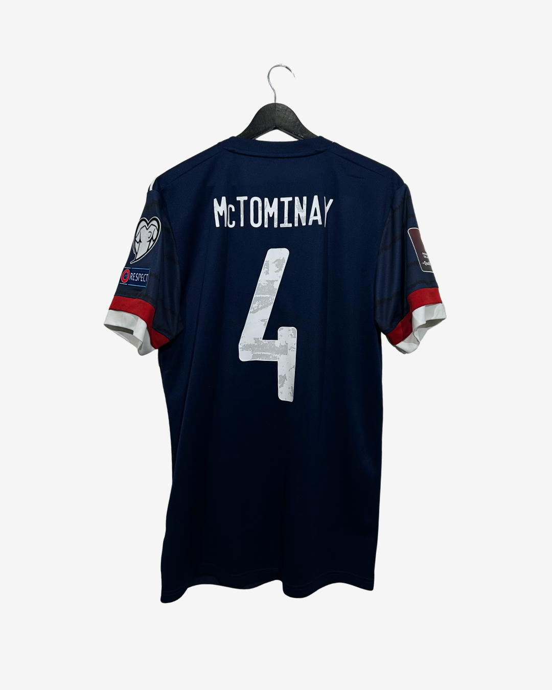 A navy blue short-sleeve soccer jersey hanging on a black hanger against a light gray background. The back of the jersey features the name 'MCTOMINAY' and the number '4' in white, with red and white sleeve accents.