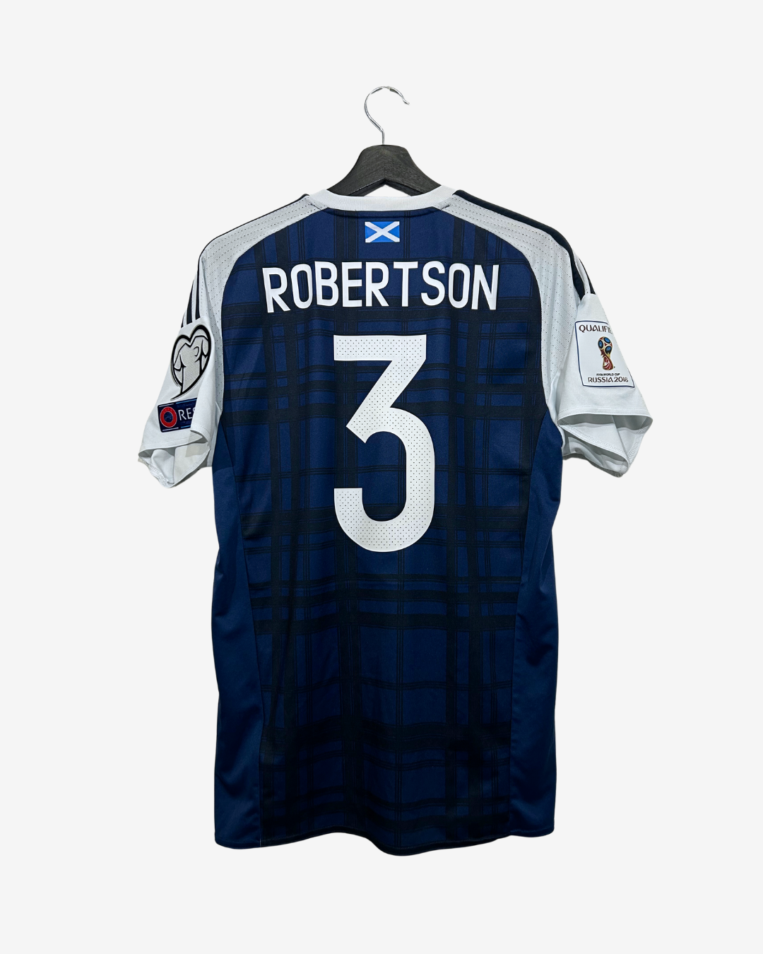 Back view of a Scotland national football team jersey featuring a dark blue plaid pattern, white shoulder panels, 'ROBERTSON' and the number '3' printed in white, with official Champions League patches on the sleeves, hanging on a black hanger against a light gray background.