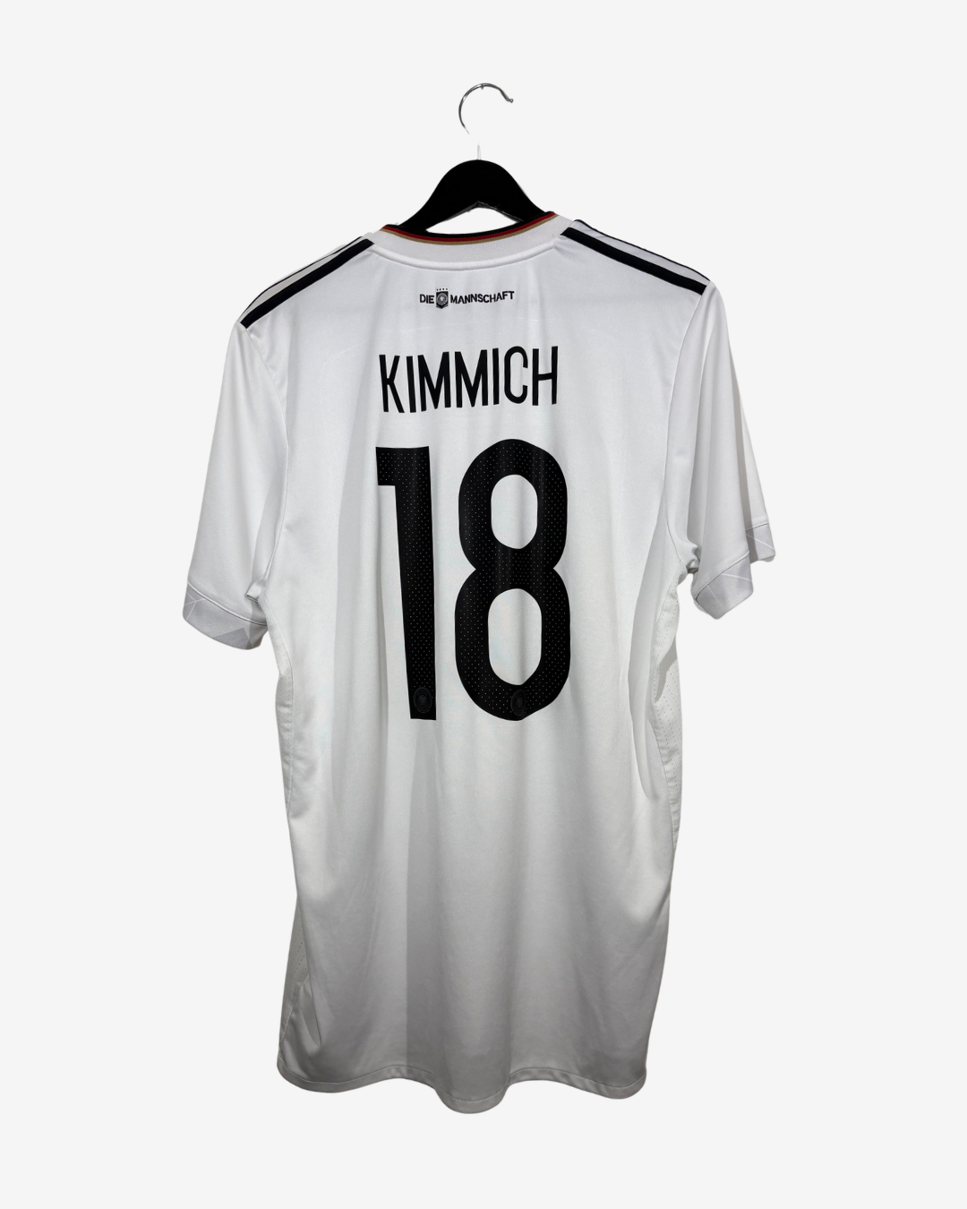 A white short-sleeve soccer jersey hanging on a black hanger against a light gray background. The back of the jersey features the name 'KIMMICH' and the number '18' in large black print, with black shoulder stripes and a small logo printed near the collar.