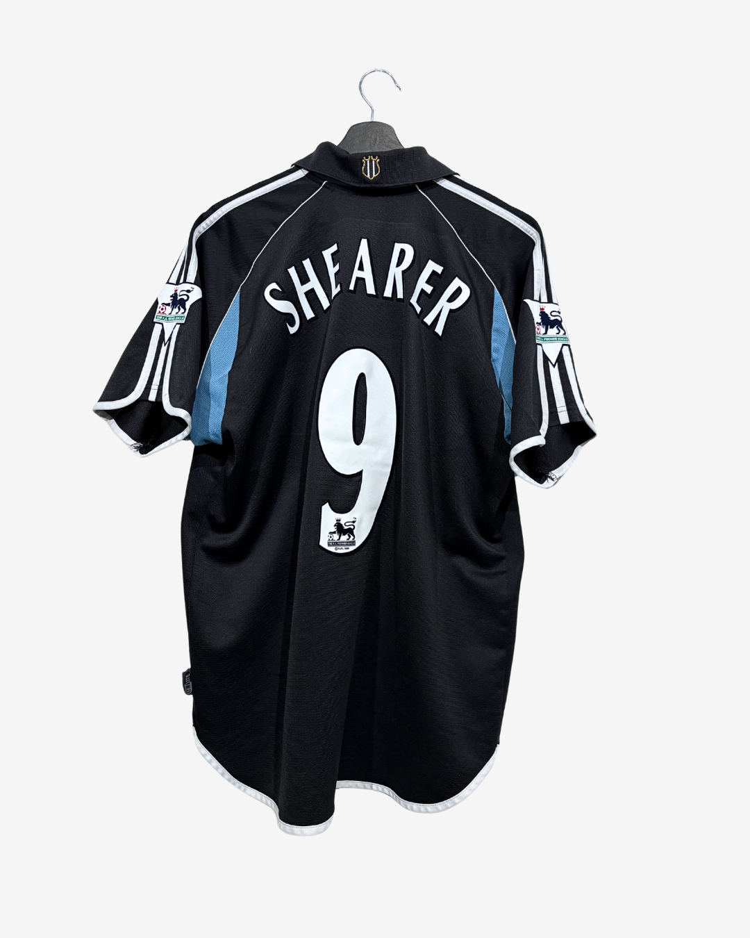 A black Newcastle United soccer jersey with 'SHEARER' and the number '9' on the back, hanging on a black hanger against a light gray background.