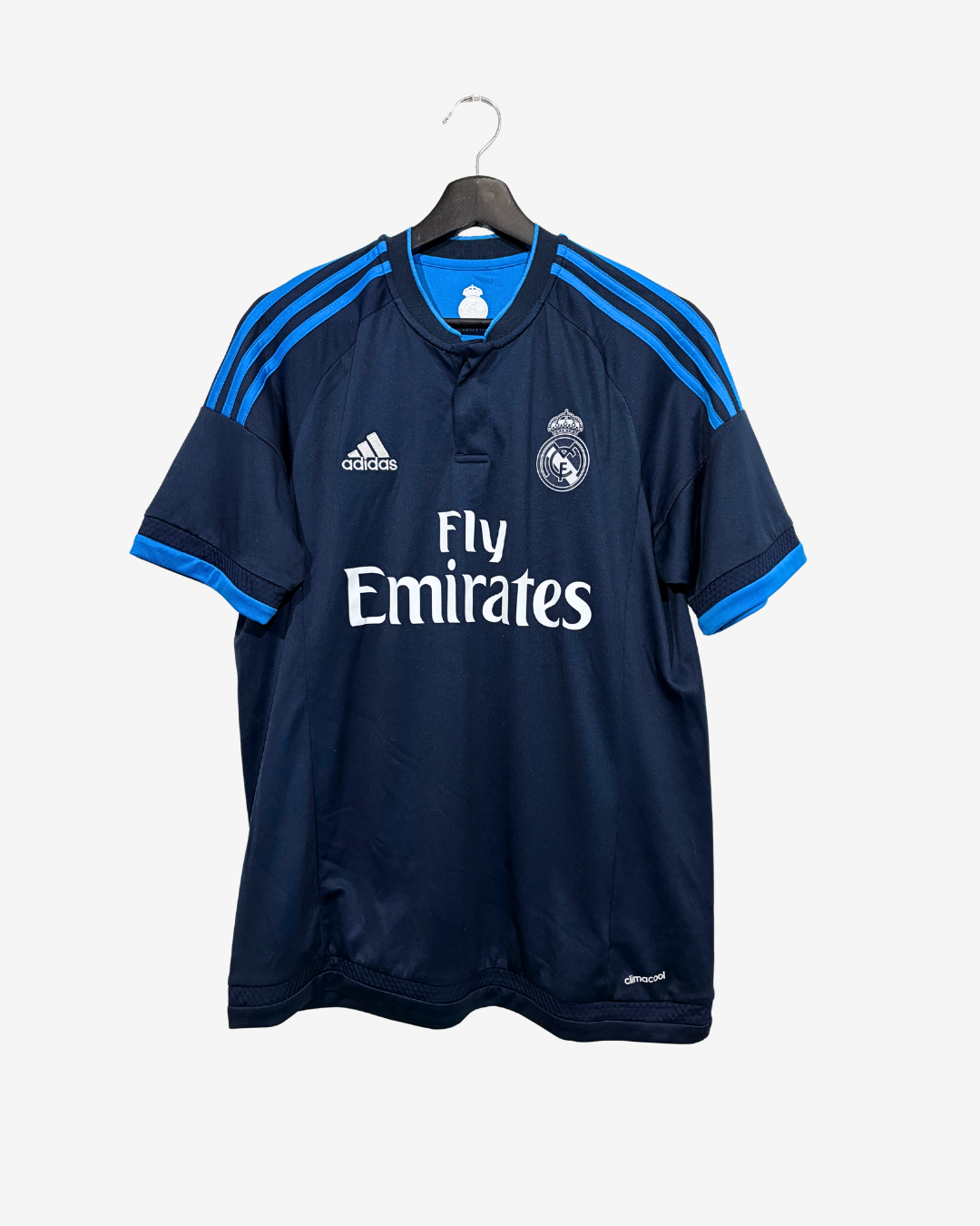 A navy blue Real Madrid soccer jersey by Adidas, featuring the white Fly Emirates sponsor logo, the club crest, and blue shoulder stripes, hanging on a black hanger against a light gray background.