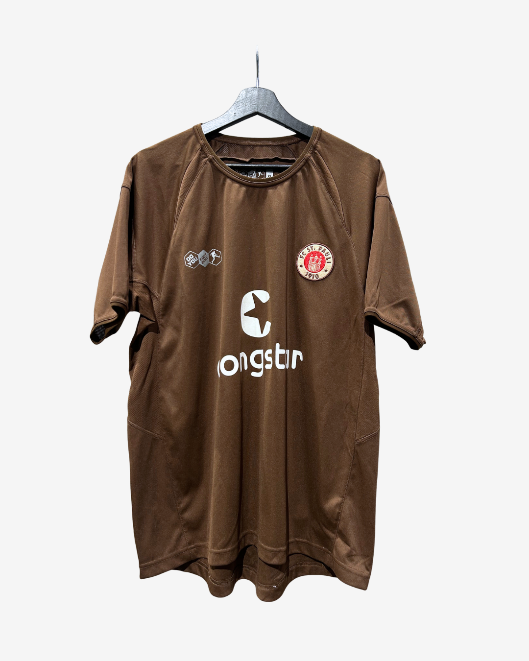 A brown short-sleeve athletic jersey with white printed logos, hanging on a gray hanger against a light gray background. The jersey features a circular red and white club crest on the left chest and a large white 'ongstin' sponsor logo in the center.