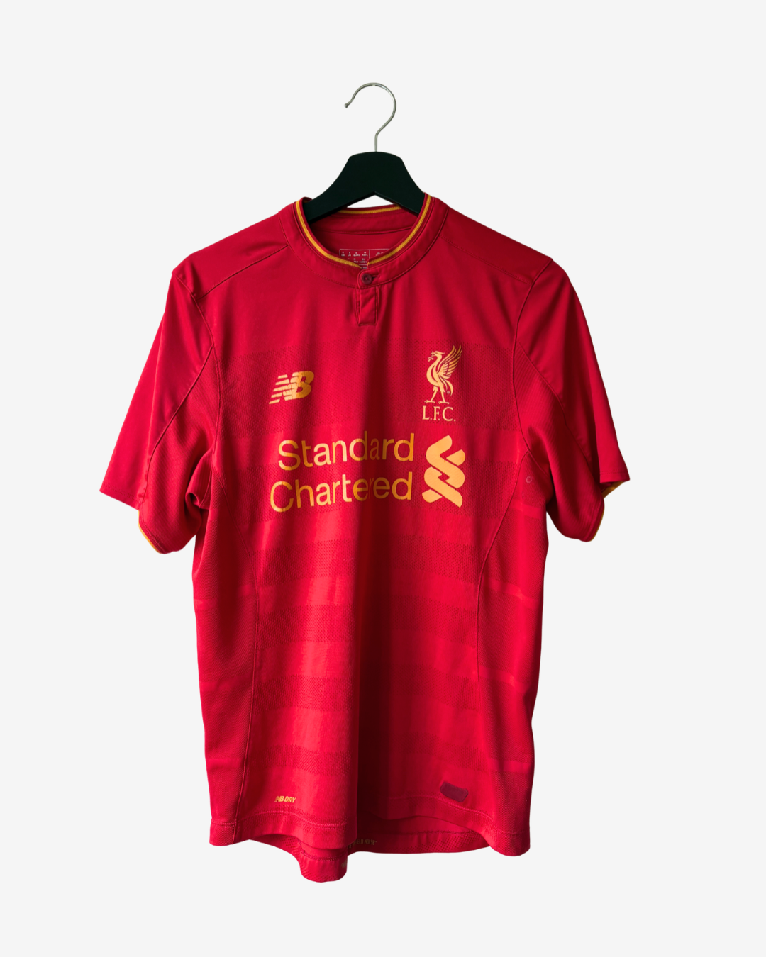 A red short-sleeve Liverpool football jersey hanging on a black hanger against a light gray background. The jersey features yellow/gold New Balance branding, the Liverpool FC crest, and 'Standard Chartered' sponsor text.