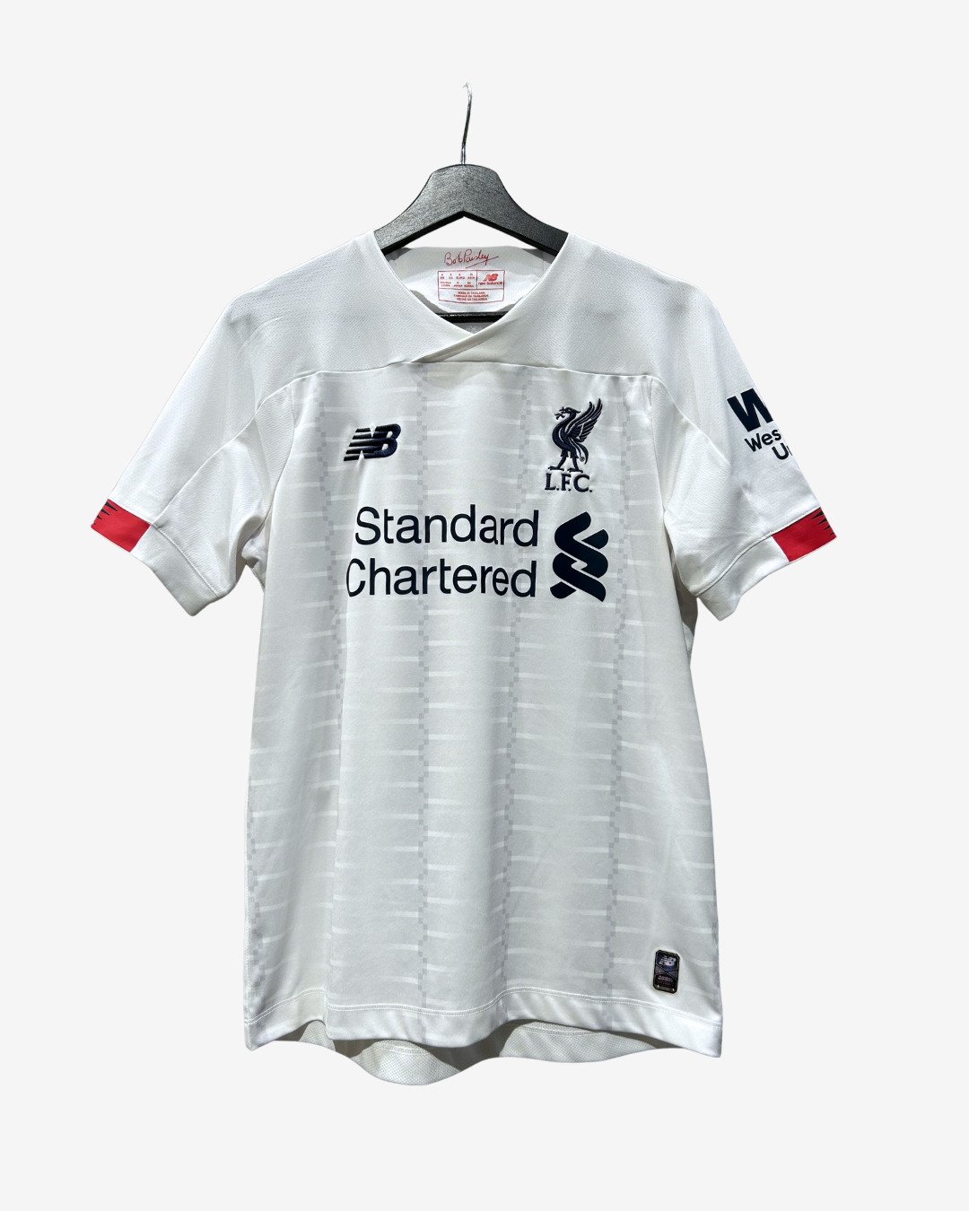 A white short-sleeve Liverpool football jersey hanging on a black hanger against a light gray background. The jersey features the black New Balance logo on the right chest, the black Liverpool F.