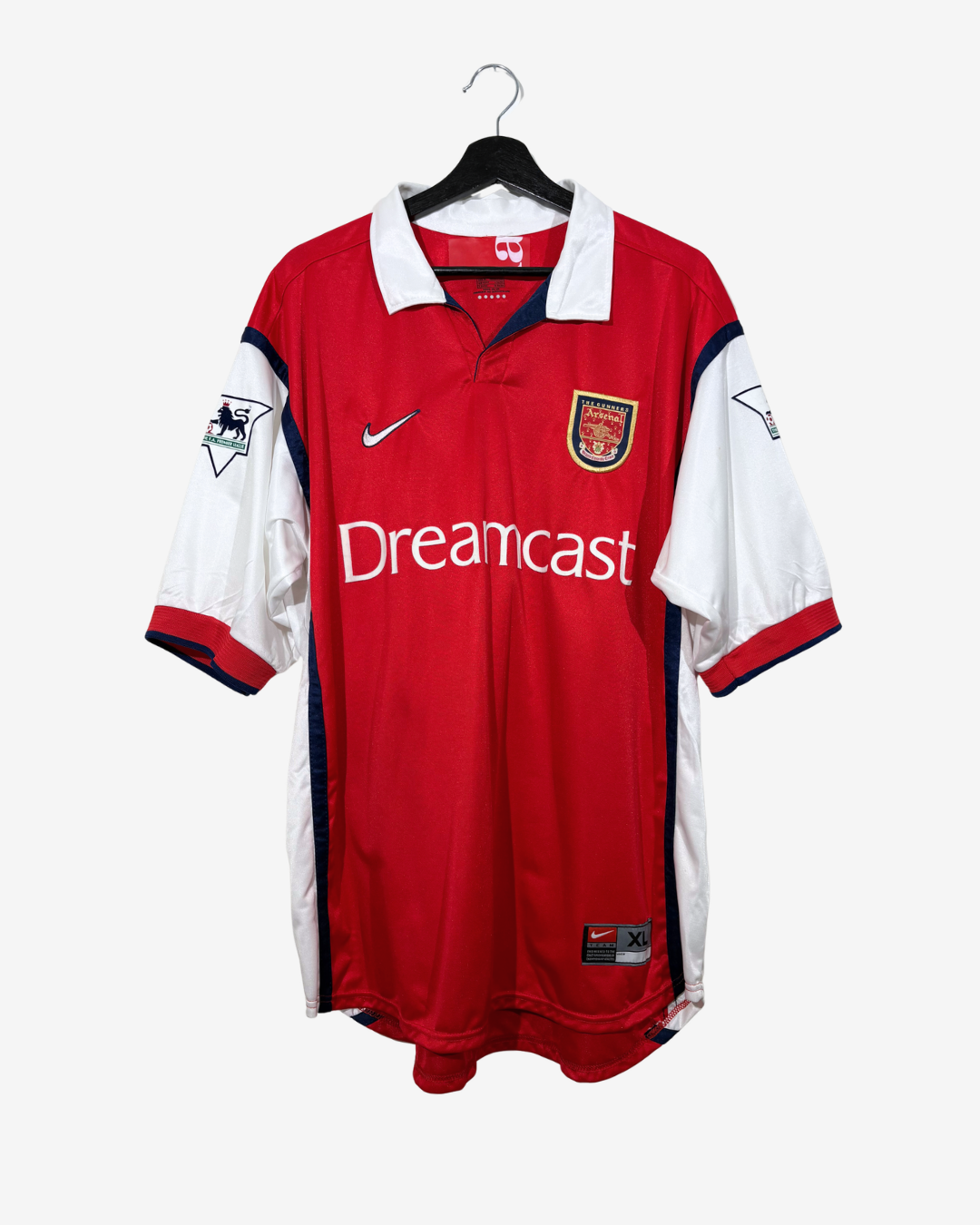 A red and white Arsenal soccer jersey hanging on a black hanger against a light gray background. The jersey features the white 'Dreamcast' sponsor logo across the chest, a white Nike swoosh on the right chest, and the Arsenal club crest on the left chest.