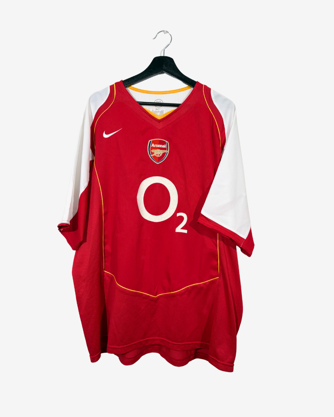 A red and white Arsenal football jersey hanging on a black hanger against a light gray background. The jersey features the Arsenal club crest on the chest, a white Nike swoosh, and a large white O2 logo.