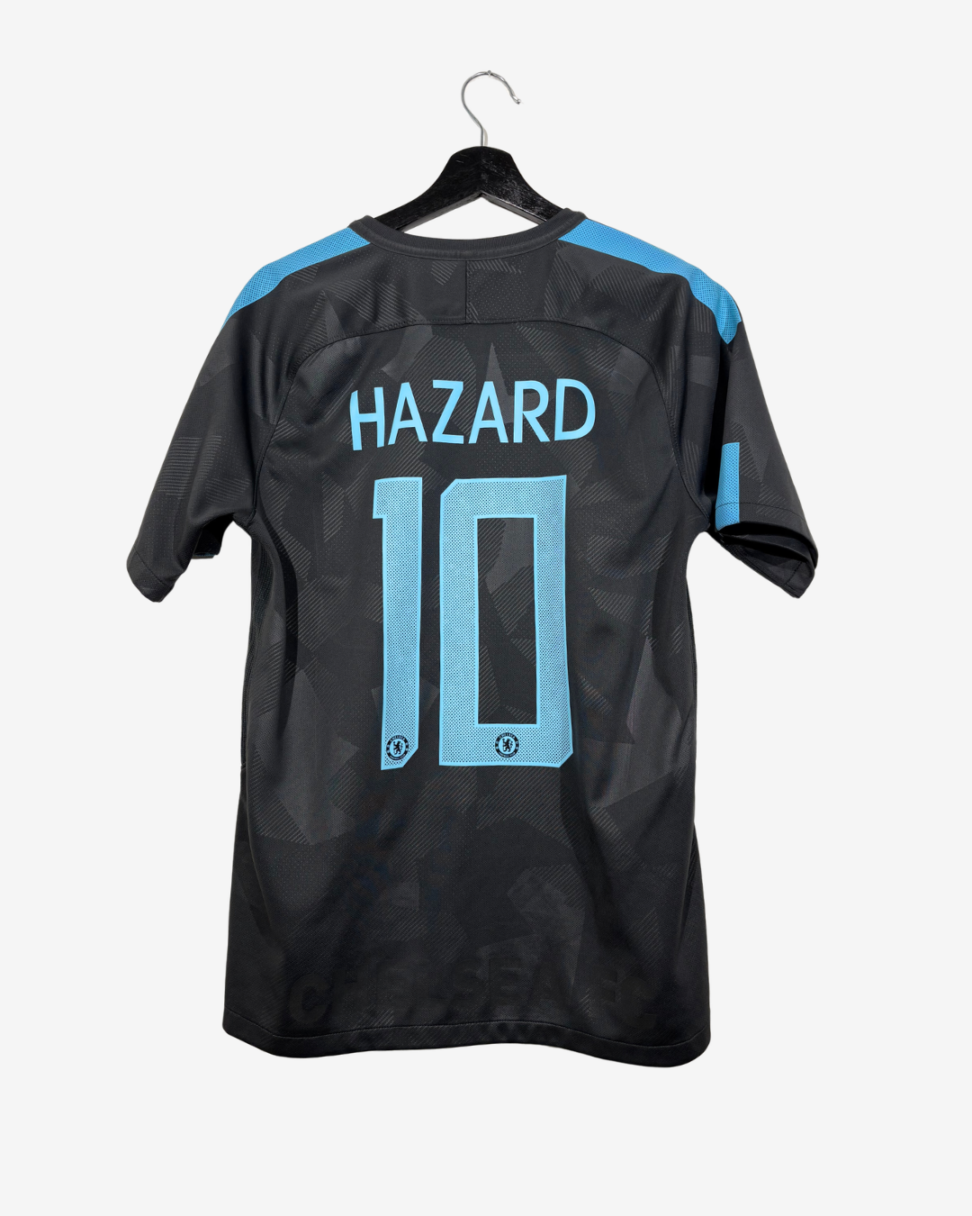 Back view of a black Chelsea FC  football jersey with light blue shoulder stripes, 'HAZARD' and number '10' printed in light blue on the back, hanging on a black hanger against a light gray background.