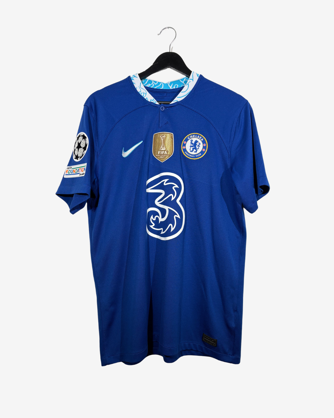 A blue Chelsea football jersey hanging on a black hanger against a light gray background. The jersey features the white Nike swoosh, the Chelsea crest, a gold UEFA Champions League patch, and the white 3 logo on the front.