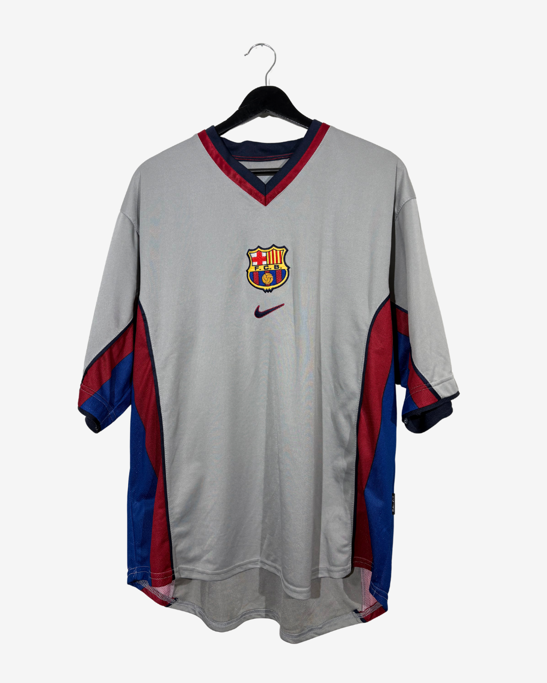 A vintage FC Barcelona home soccer jersey by Nike, featuring a light gray body with red and blue side panels and a red and blue V-neck collar, hanging on a black hanger against a light gray background.
