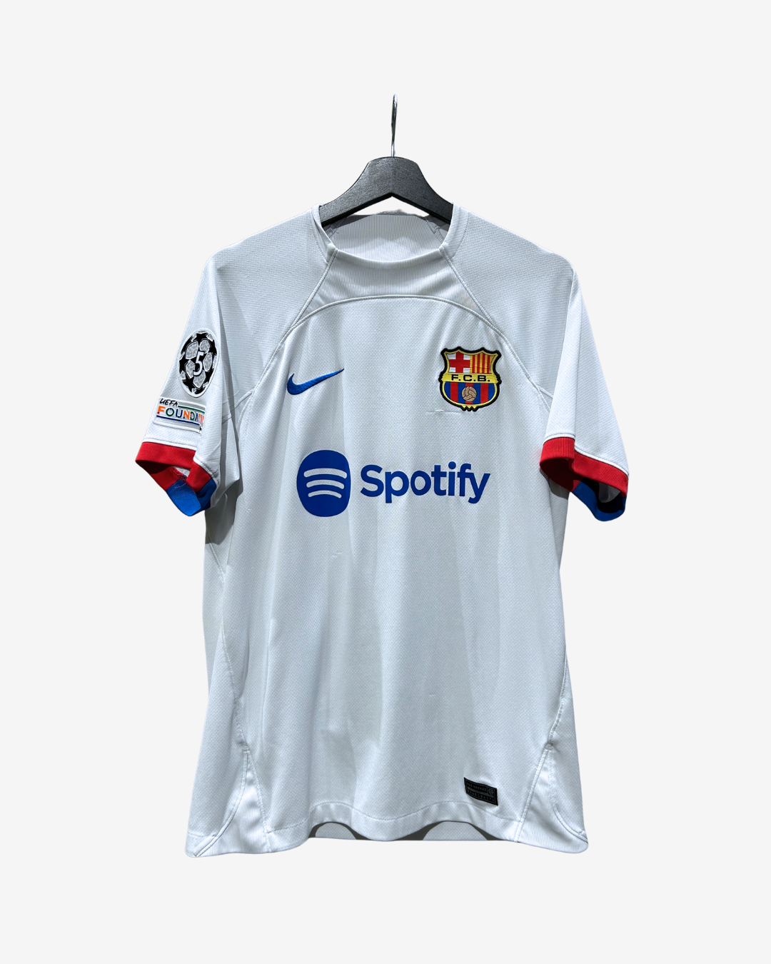 A white FC Barcelona home soccer jersey from the 2015-2016 season, featuring the blue Nike swoosh and Spotify sponsor logo on the front, hanging on a black hanger against a light gray background.