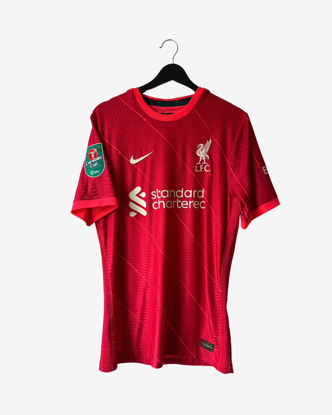 A red Liverpool FC Nike soccer jersey hanging on a black hanger against a light gray background. The jersey features the white Nike swoosh, the Liverpool FC crest, the 'standard chartered' sponsor logo, and a green 2023 Champions League patch on the sleeve.