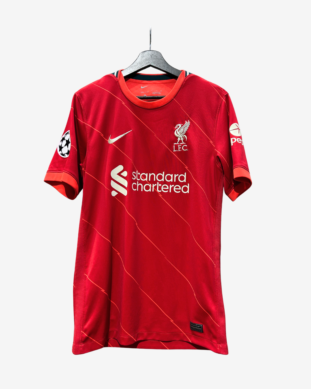 A red Liverpool football jersey hanging on a black hanger against a light gray background. The jersey features the white Nike swoosh, the Liverpool FC crest, the Standard Chartered sponsor logo, and a UEFA Champions League patch on the sleeve.