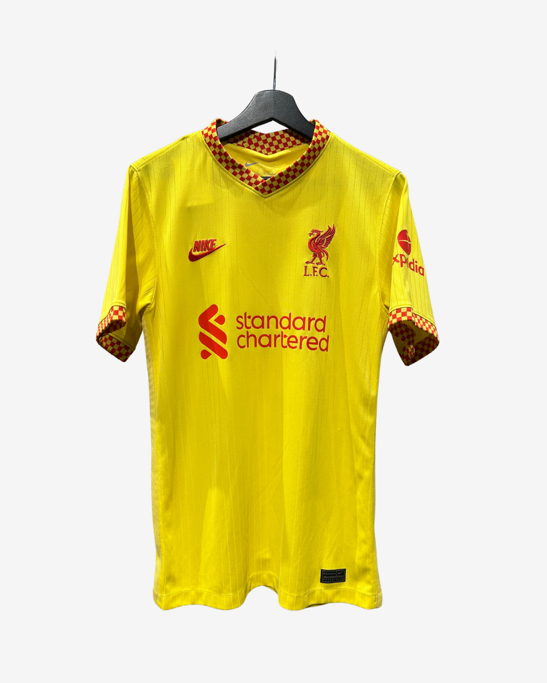 A yellow Liverpool football jersey with red and brown checkered trim on the collar and cuffs, featuring the Nike swoosh, LFC crest, and Standard Chartered sponsor logo, hanging on a black hanger against a light gray background.