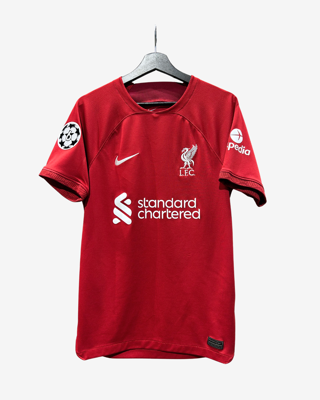 A red Liverpool FC short-sleeve soccer jersey hanging on a black hanger against a light gray background. The jersey features the white Nike swoosh, the Liverpool FC crest, the UEFA Champions League patch on the right sleeve, and the 'standard chartered' sponsor logo across the center chest, with the 'pedra' sponsor logo on the left sleeve.