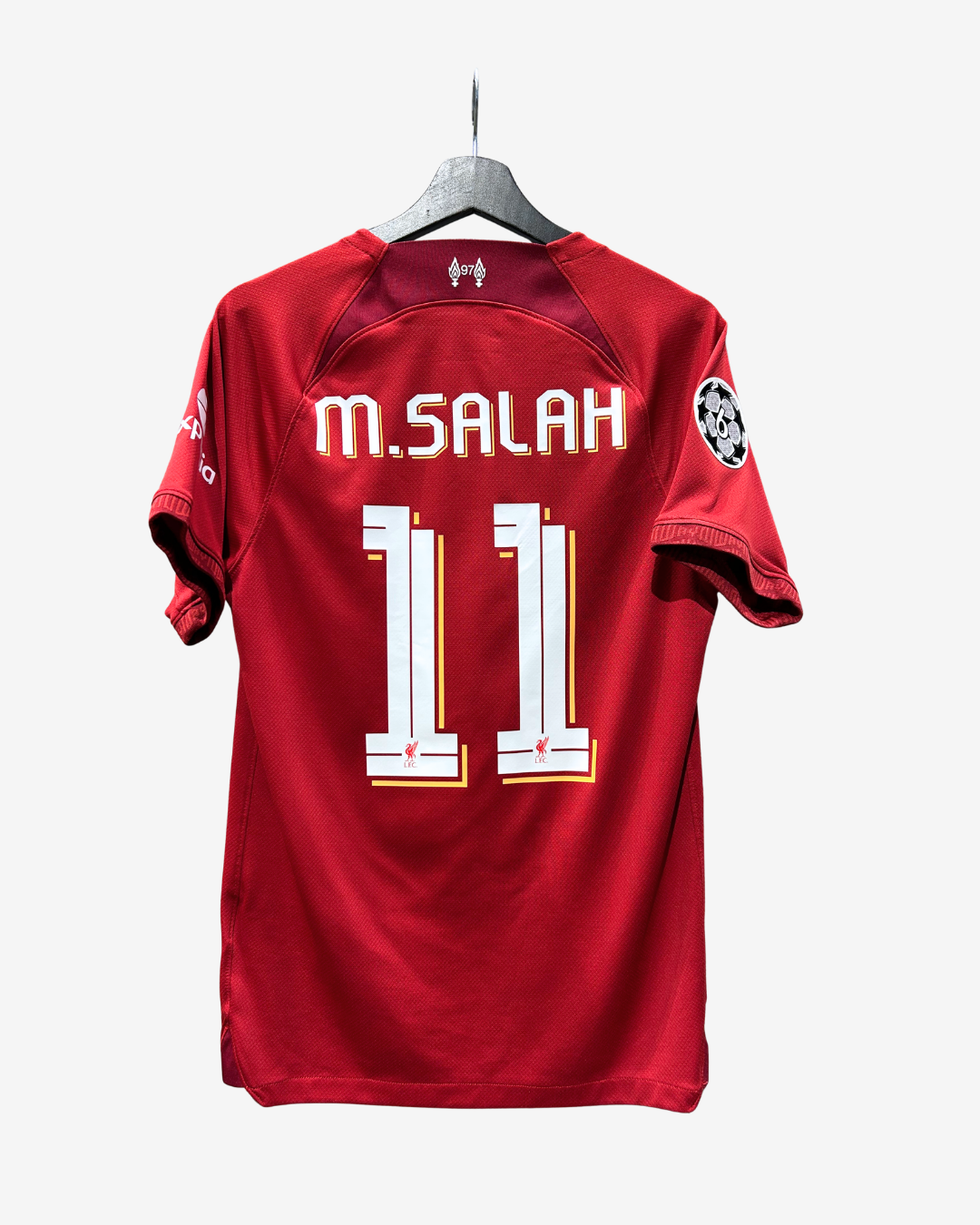 A red Under Armour sports jersey with 'M. SALAH' and the number '11' printed on the back, hanging on a gray hanger against a light gray background.