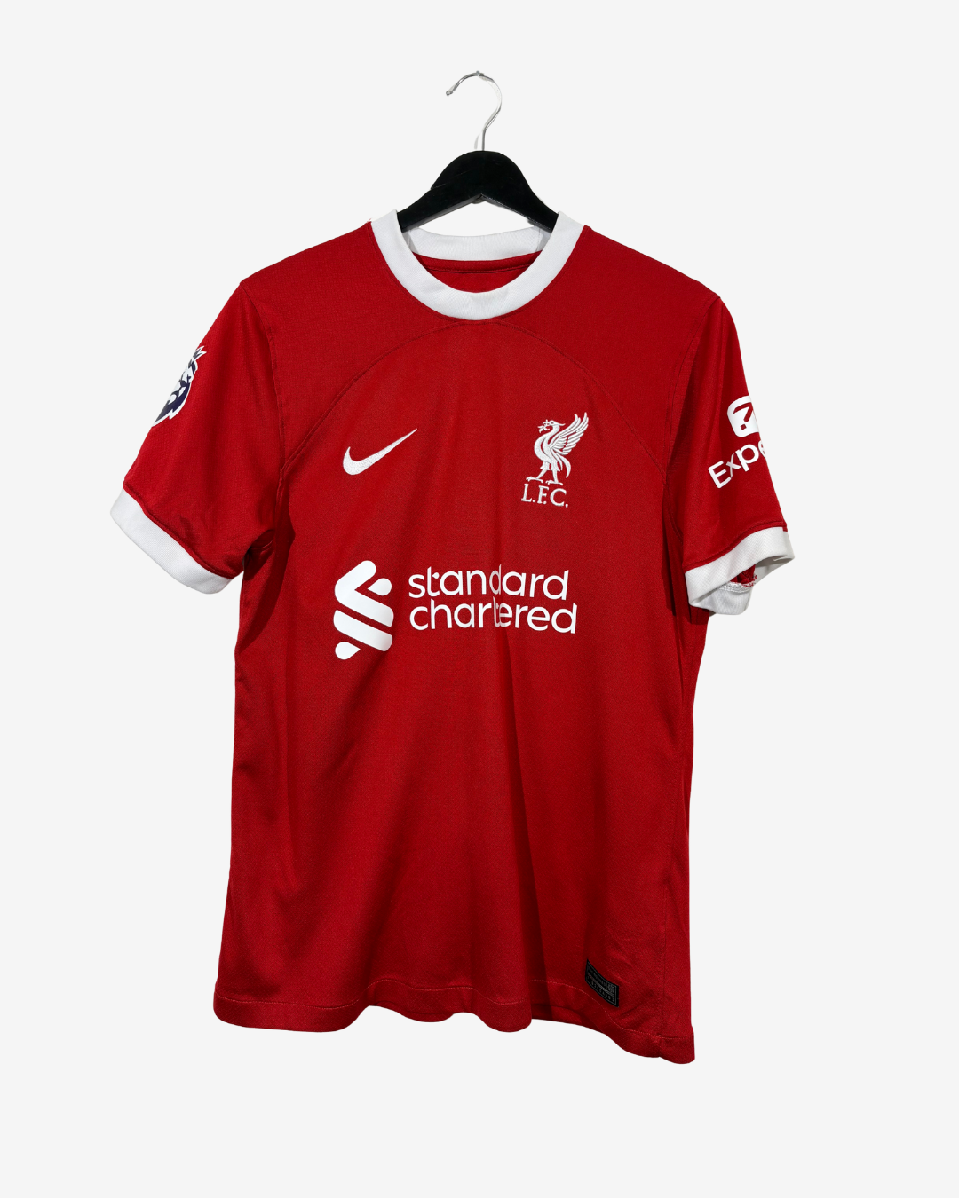 A red Liverpool FC short-sleeve soccer jersey by Nike, featuring white trim on the collar and cuffs, with the white 'standard chartered' sponsor logo across the center chest, displayed on a black hanger against a light gray background.