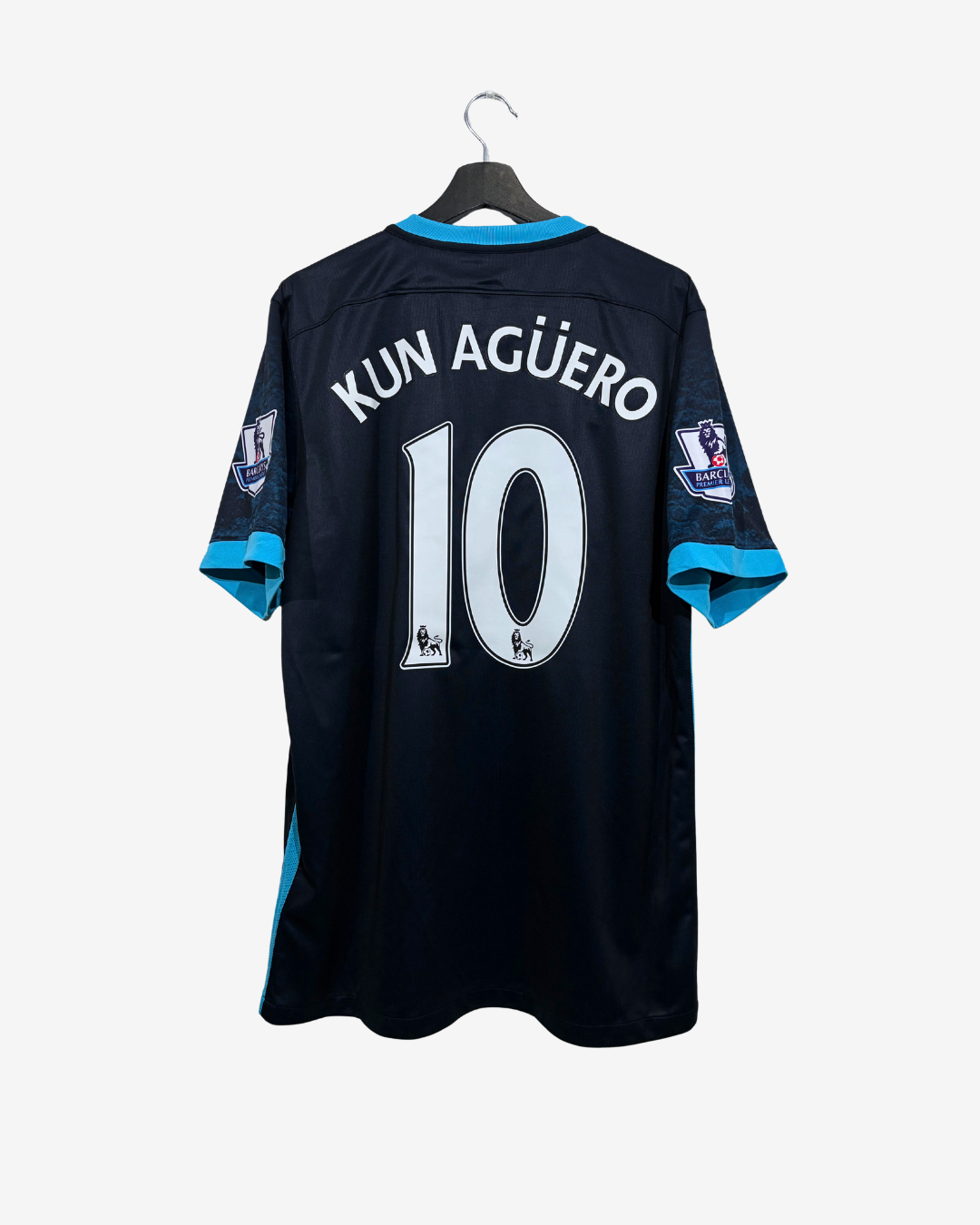 Back view of a dark navy blue soccer jersey hanging on a black hanger against a light gray background. The jersey features the name 'KUN AGÜERO' and the number '10' in white with a light blue outline, and light blue trim on the collar and sleeve cuffs.