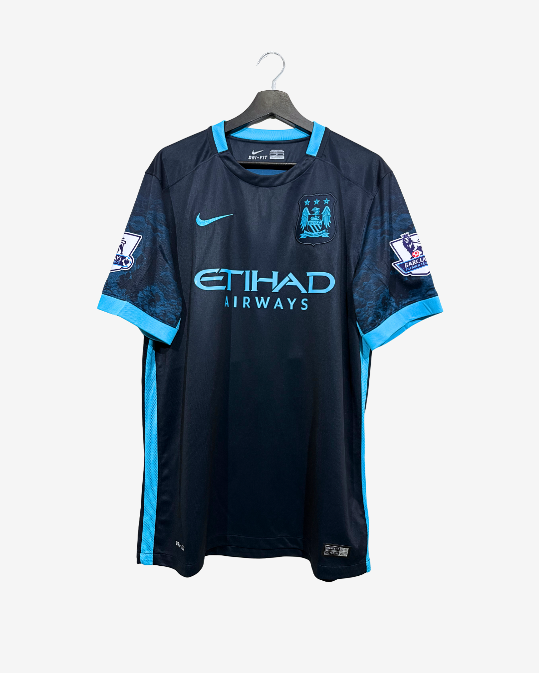 A Manchester City home soccer jersey by Nike, featuring a dark navy blue body with bright blue accents on the collar, cuffs, and side panels, hanging on a black hanger against a light gray background.