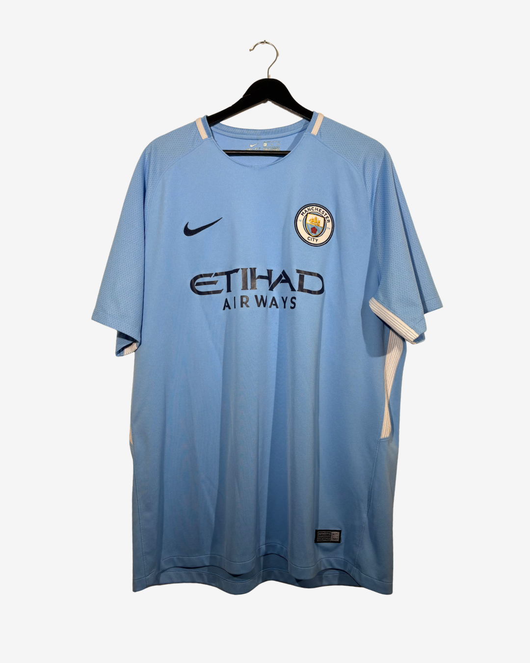 A light blue Manchester City home soccer jersey hanging on a black hanger against a light gray background. The jersey features the black Nike swoosh on the right chest, the Manchester City crest on the left chest, and 'ETIHAD AIR WAYS' printed across the center.