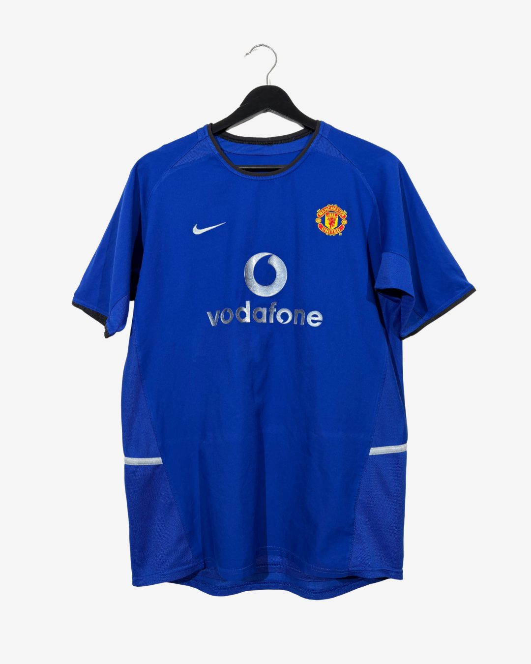 A blue Manchester United Nike soccer jersey hanging on a black hanger against a light gray background. The jersey features the white Vodafone sponsor logo and the Manchester United crest on the chest.