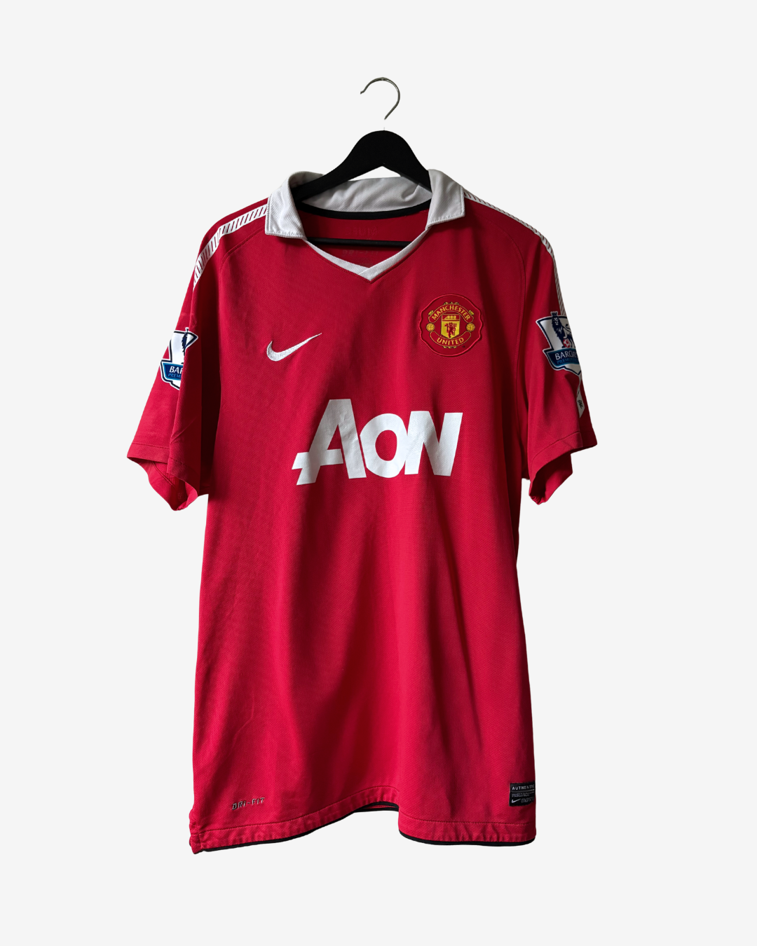 A red Manchester United Nike soccer jersey with a white AON sponsor logo and white Nike swoosh, hanging on a black hanger against a light gray background.