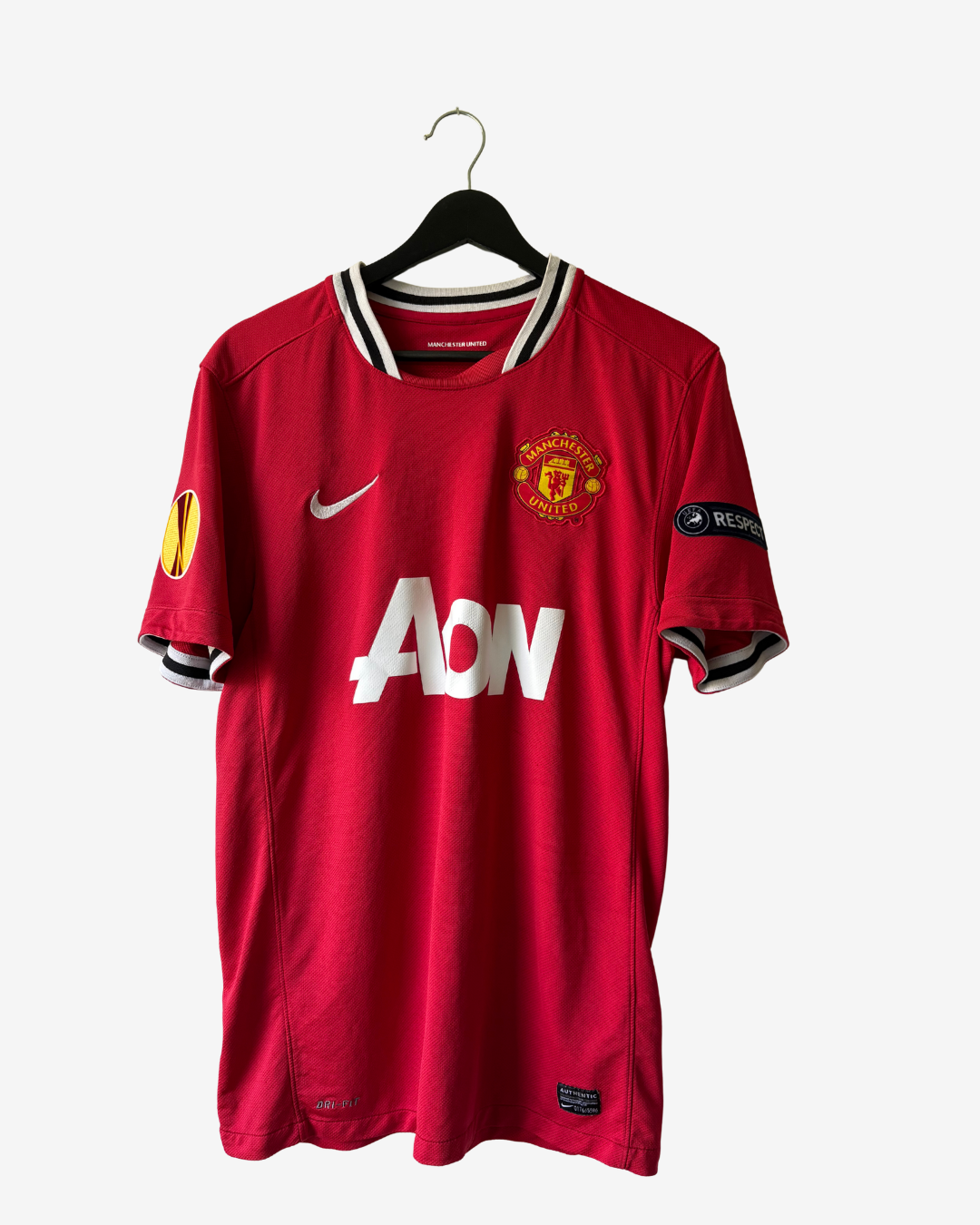 A red Manchester United Nike soccer jersey hanging on a black hanger against a light gray background. The jersey features the white AON sponsor logo on the front, the Manchester United crest on the left chest, and a Nike swoosh on the right chest.