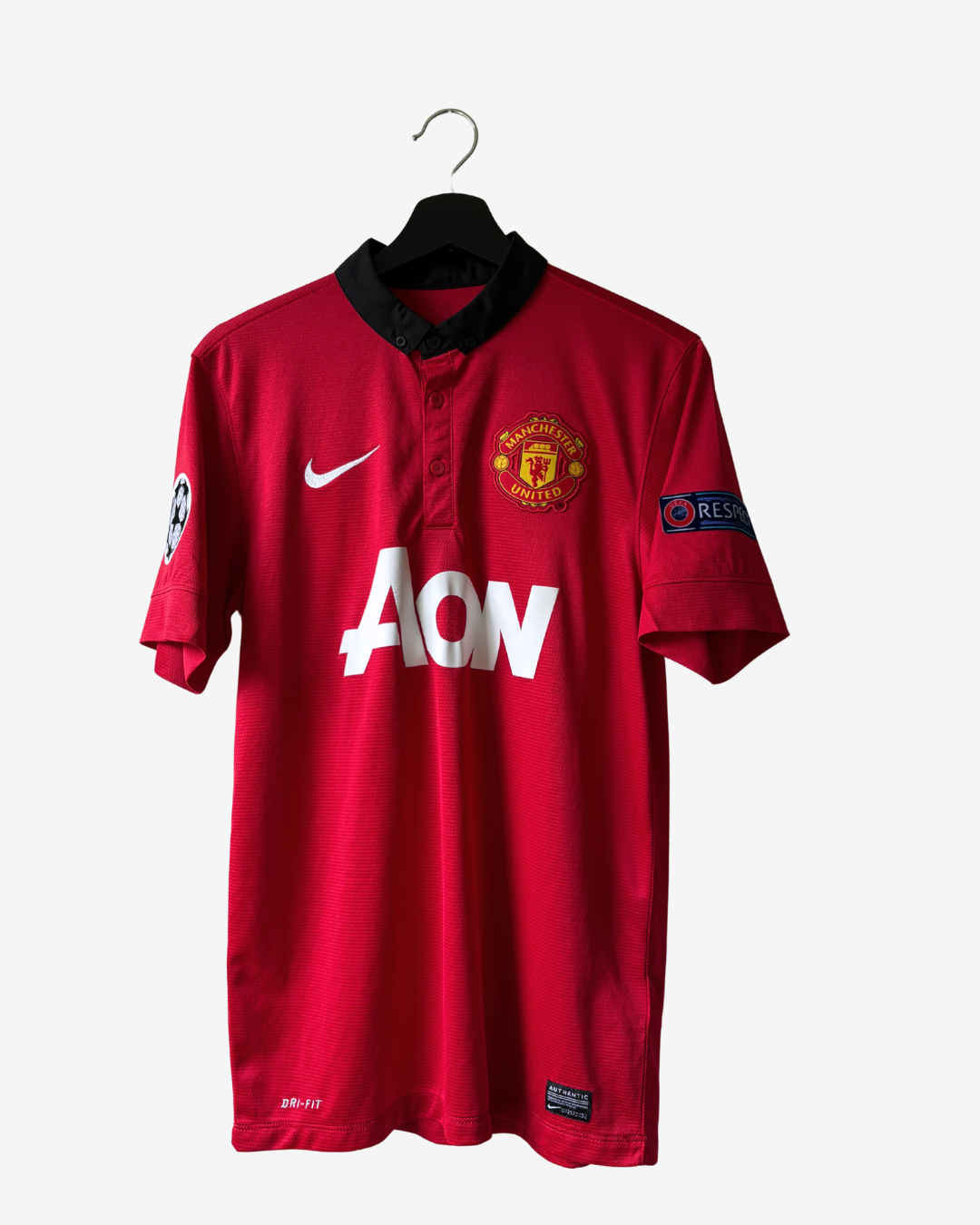 A red Manchester United Nike soccer jersey hanging on a black hanger against a light gray background. The jersey features a black collar, white AON sponsor logo, Manchester United crest, and a Nike swoosh.