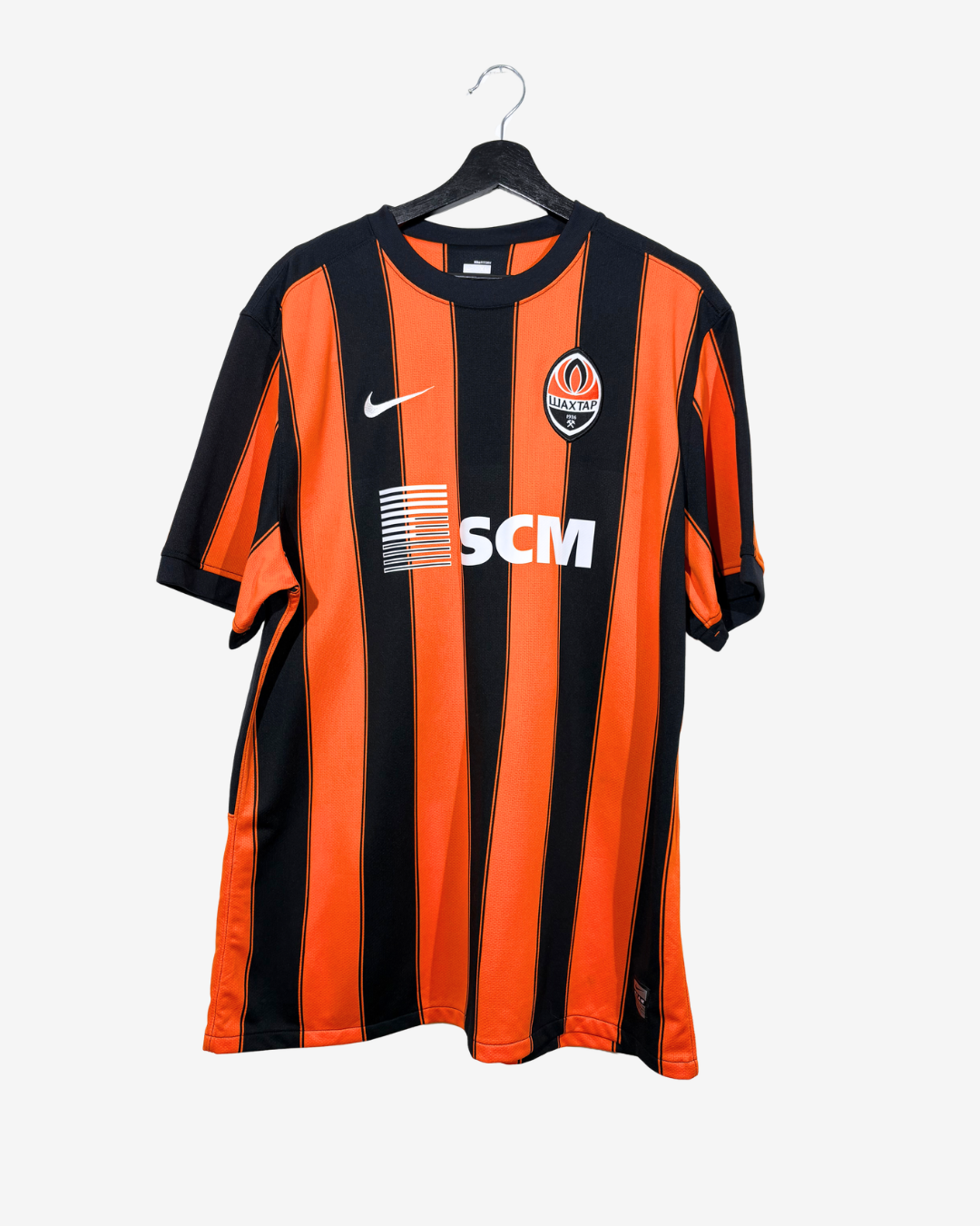 A vintage Shaktar Donetsk soccer jersey with vertical orange and black stripes, featuring a white Nike swoosh and a white SCM sponsor logo on the chest, hanging on a black hanger against a light gray background.