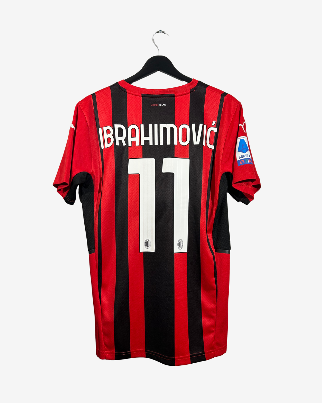 AC Milan home soccer jersey with red and black vertical stripes, featuring the name 'IBRAHIMOVIC' and number '11' on the back, hanging on a black hanger against a light gray background.