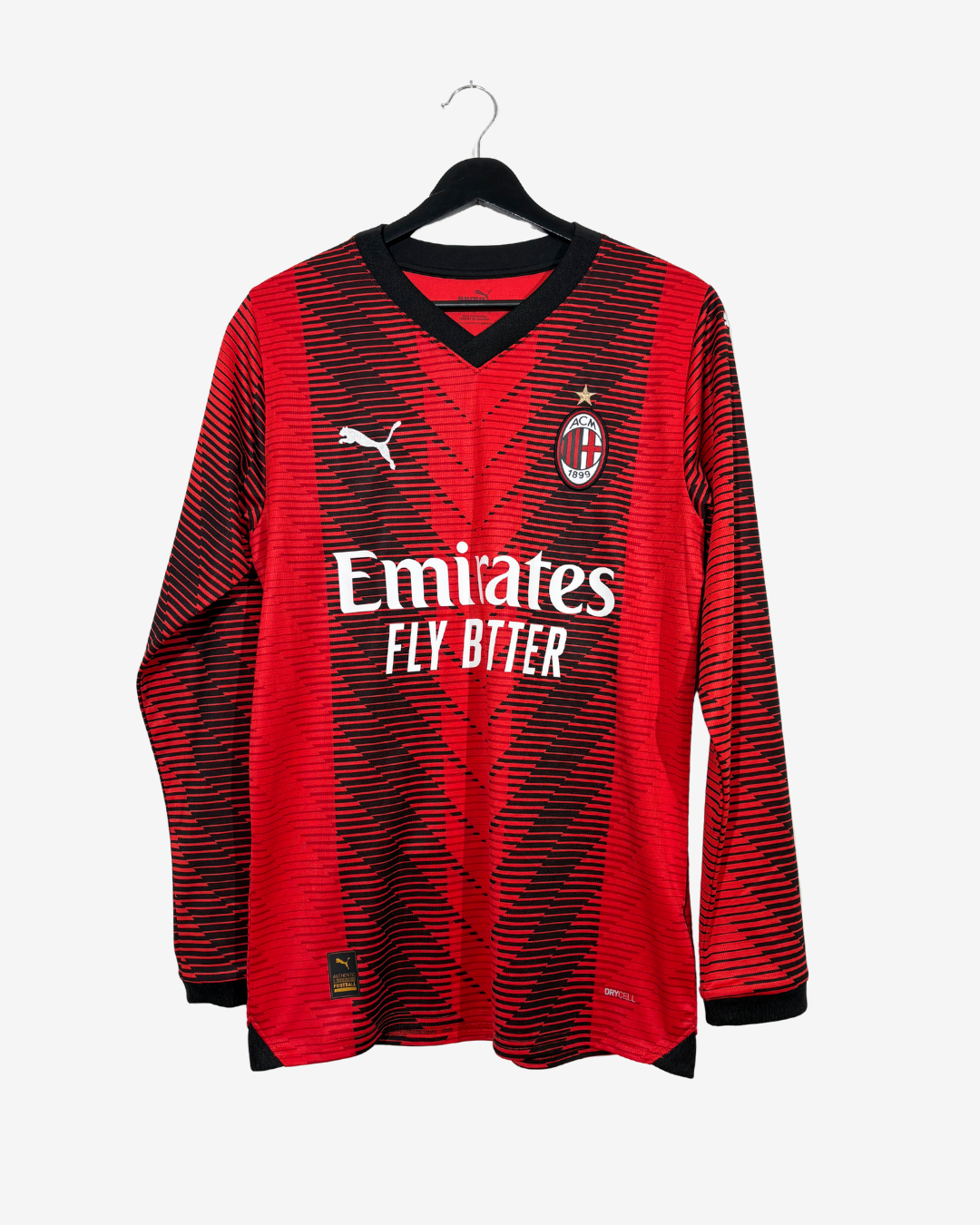 AC Milan long-sleeve soccer jersey in red and black with a subtle striped pattern, featuring the Puma logo, AC Milan crest, and Emirates Fly Better sponsor text, hanging on a black hanger against a light gray background.